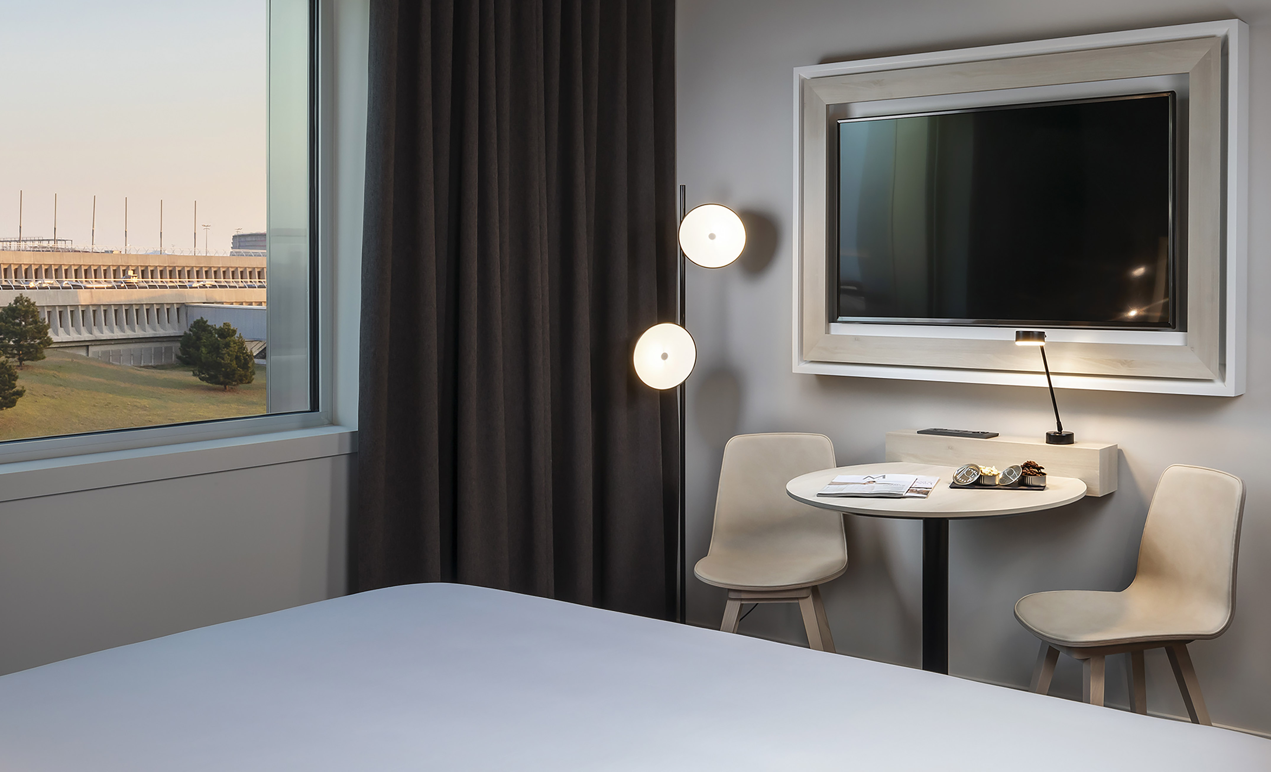 Hotel room with window view of exterior, wall-mounted TV, table, and chairs.