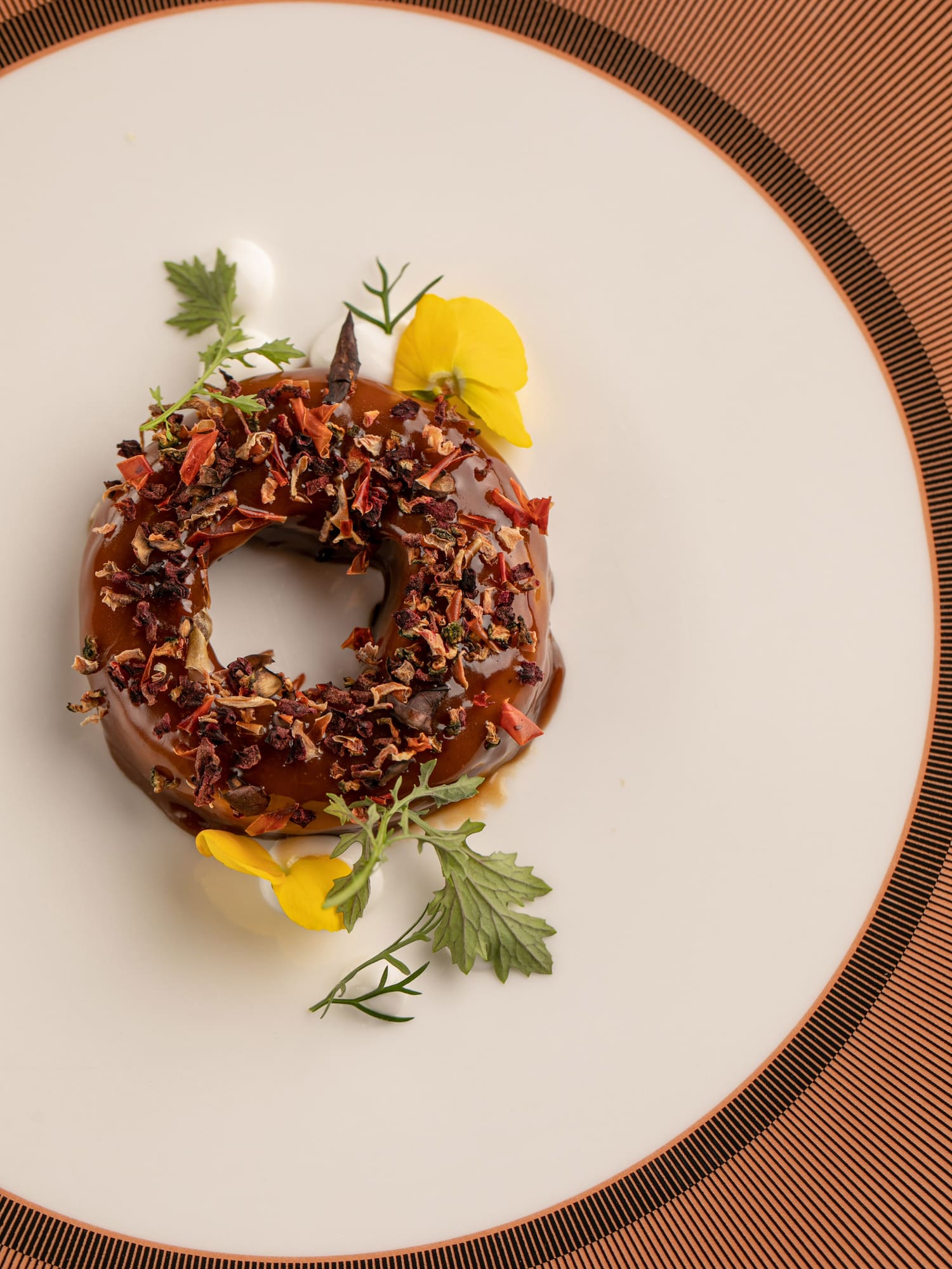 Gourmet ring dessert with brown glaze, dried topping, green herbs, and yellow flowers.