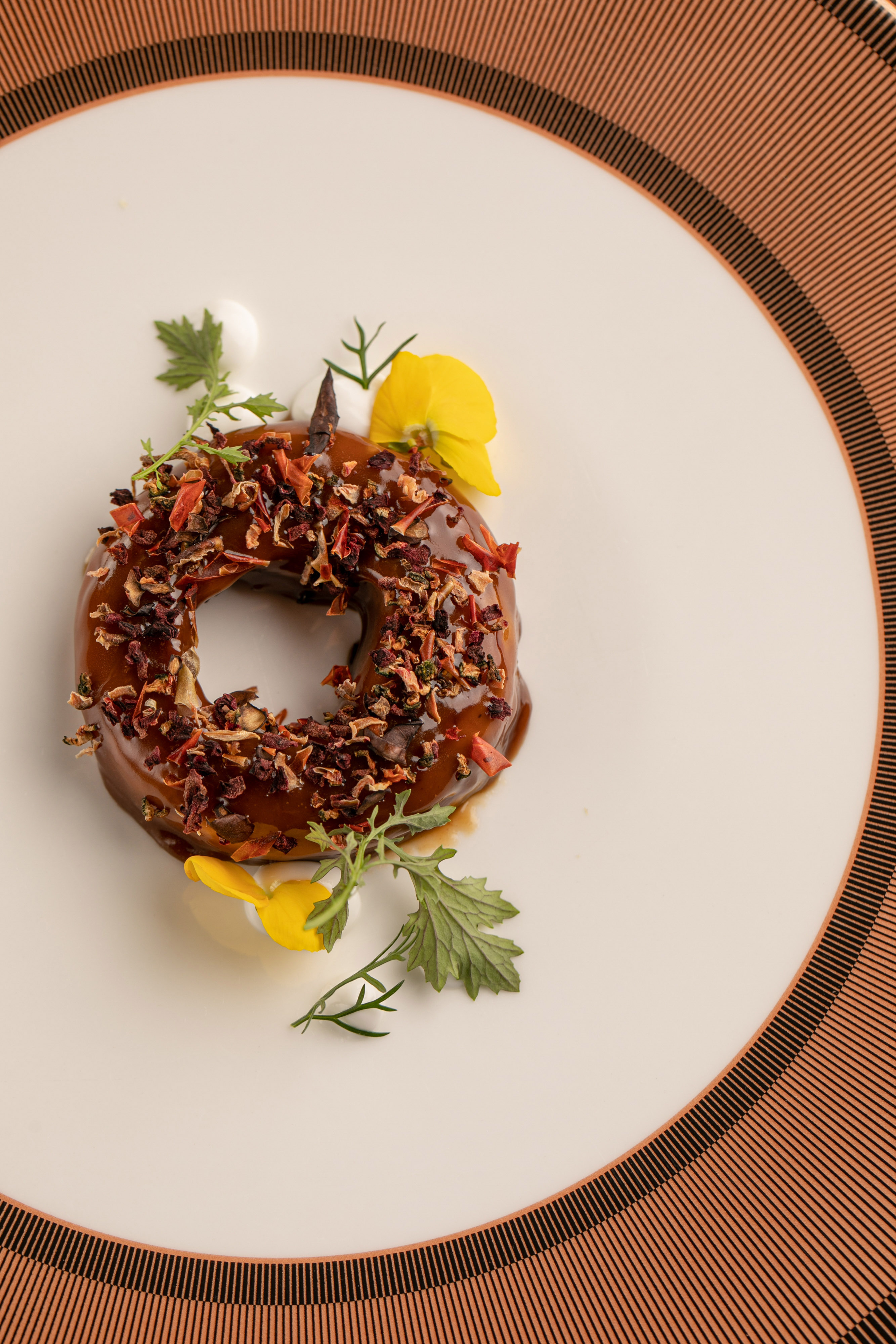 Gourmet ring dessert with brown glaze, dried topping, green herbs, and yellow flowers.