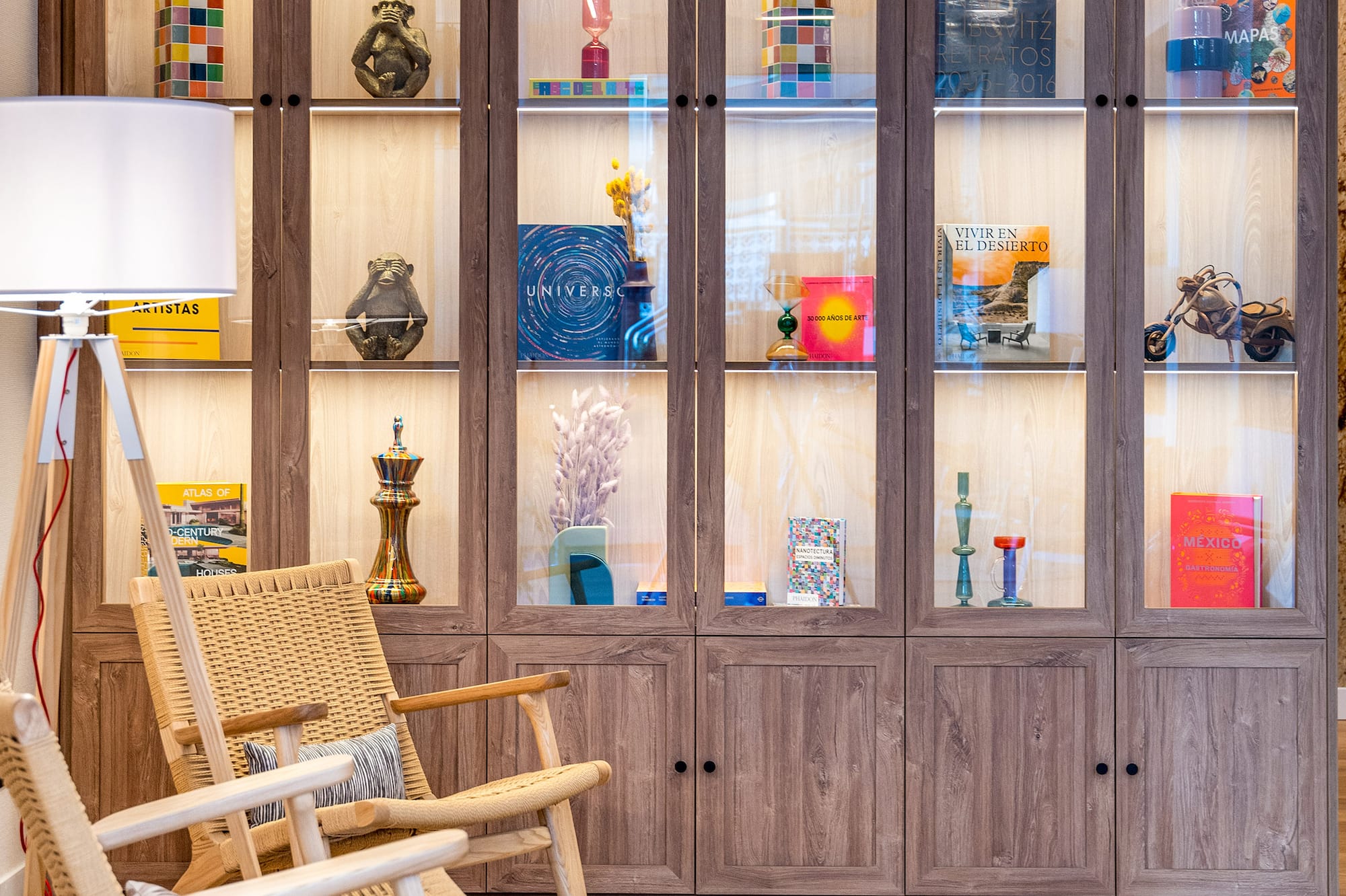 Illuminated wooden display cabinet filled with art books, decor, and rattan chairs.