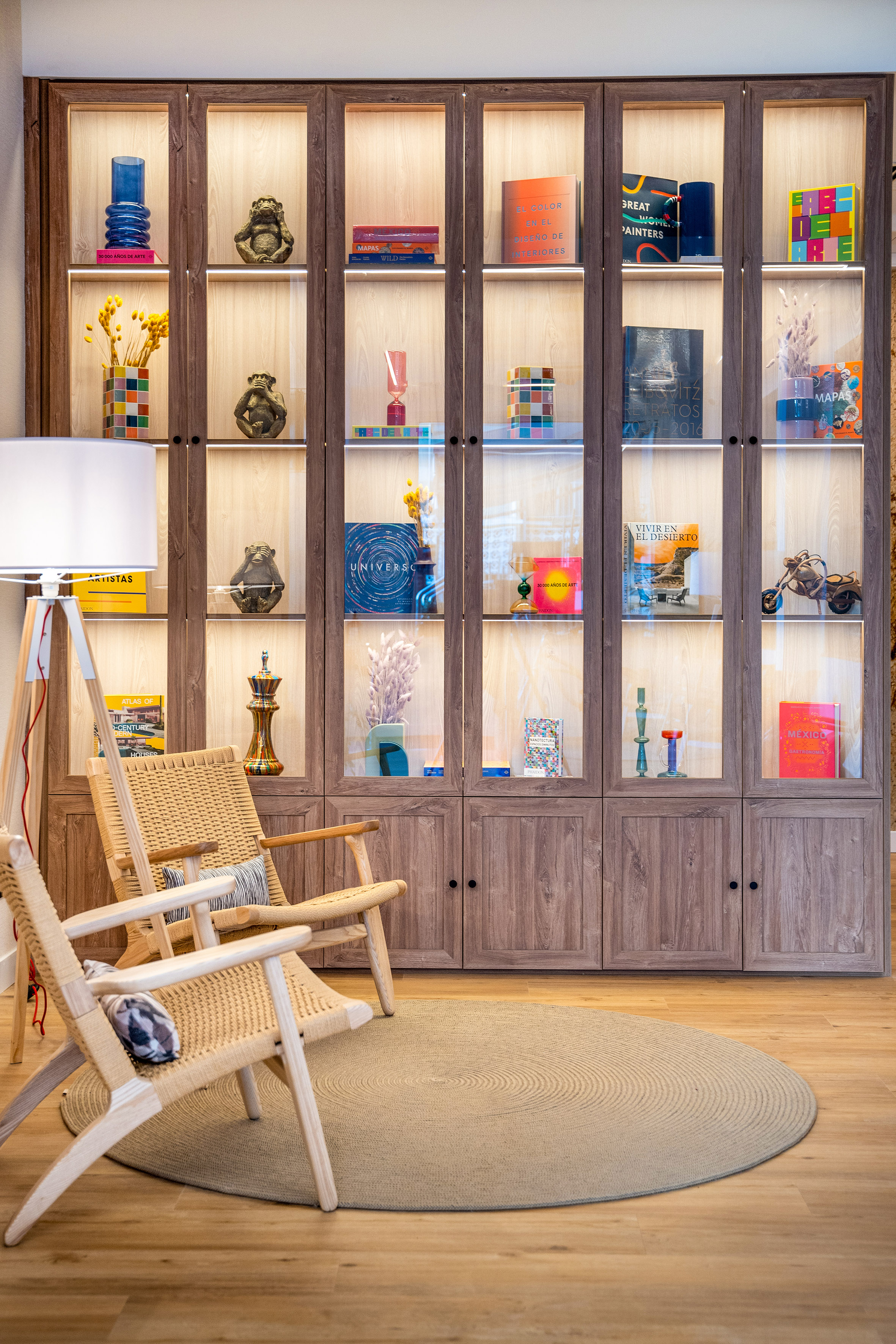 Illuminated wooden display cabinet filled with art books, decor, and rattan chairs.