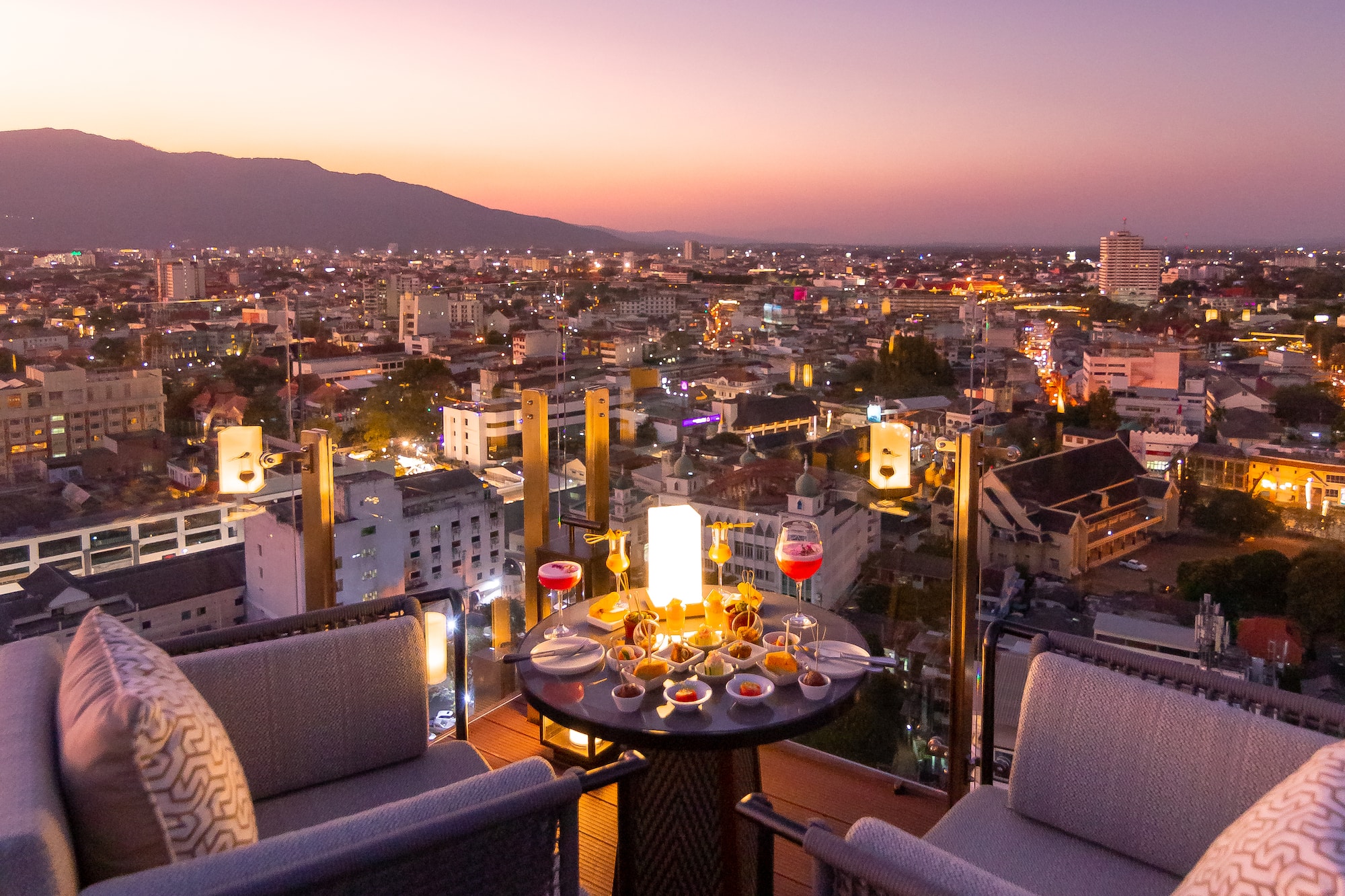 Romantic rooftop dinner overlooking city lights and mountains at sunset.