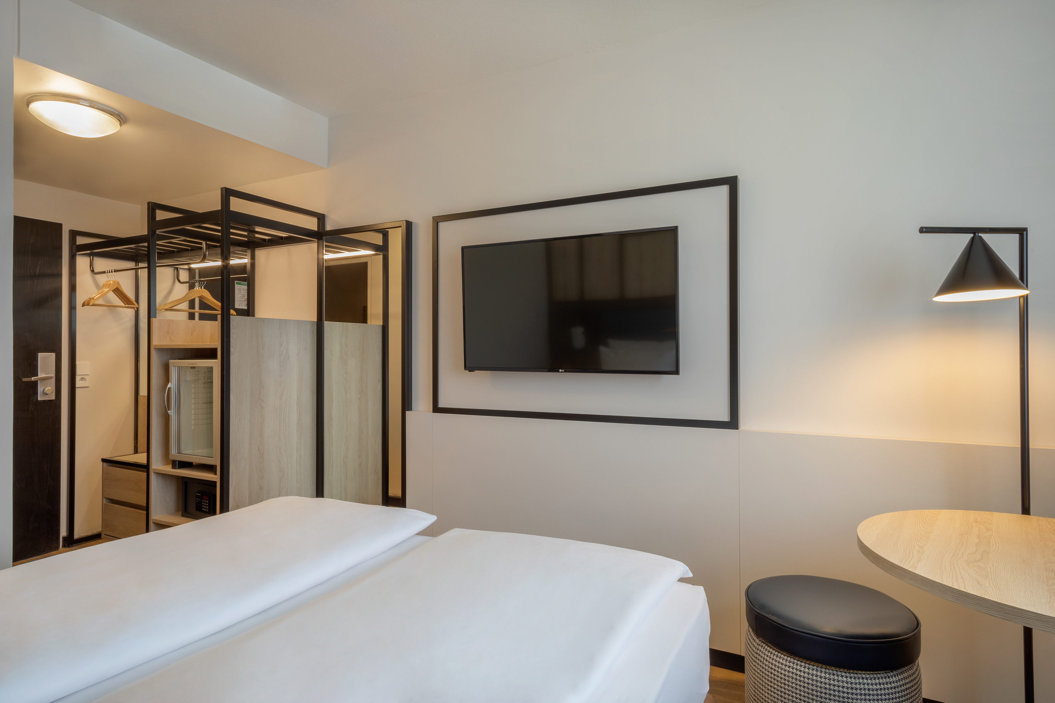 Modern hotel room with twin beds, wall TV, open wardrobe, and mini-fridge.