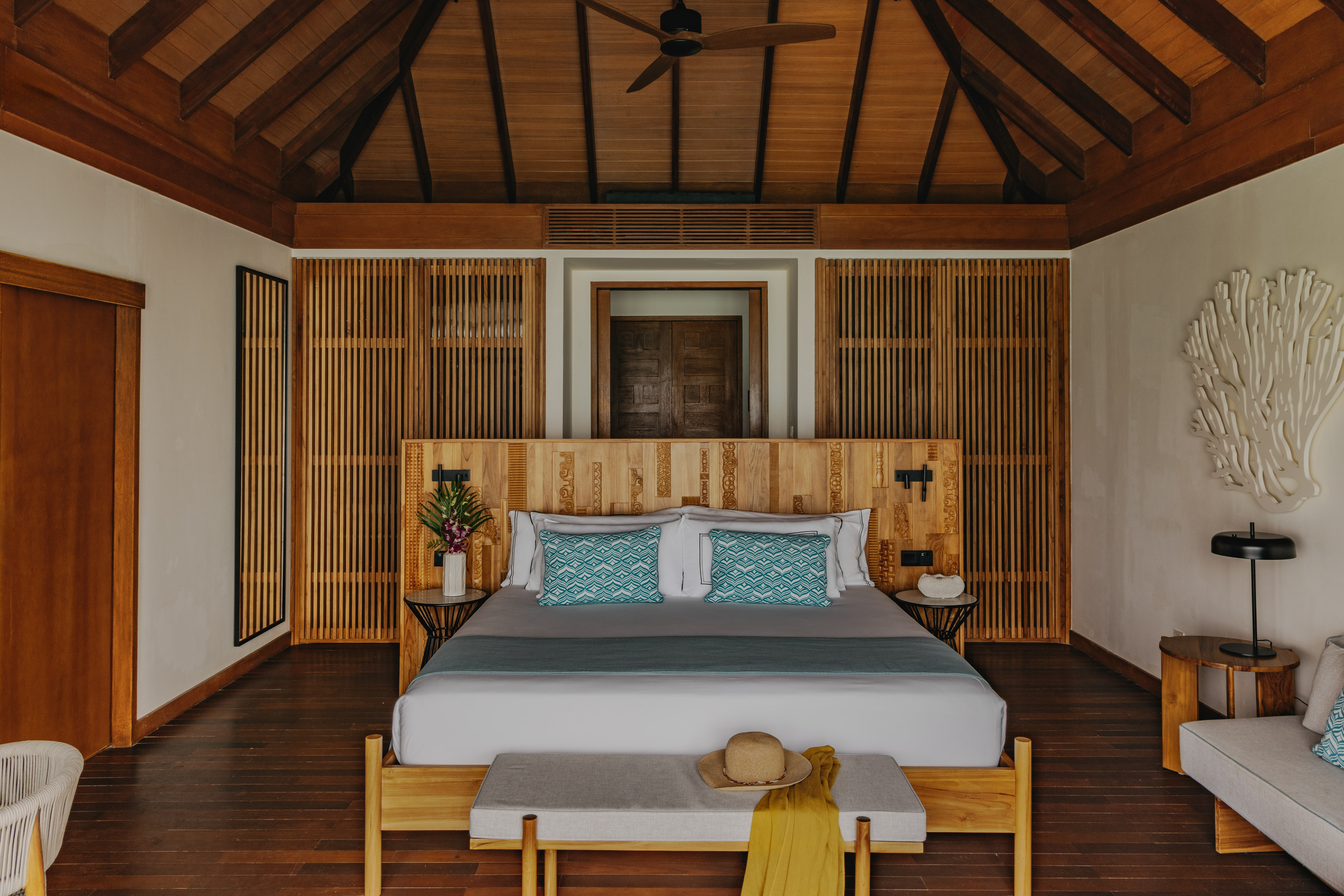 Tropical hotel bedroom with vaulted wooden ceiling, carved headboard, dark floor.