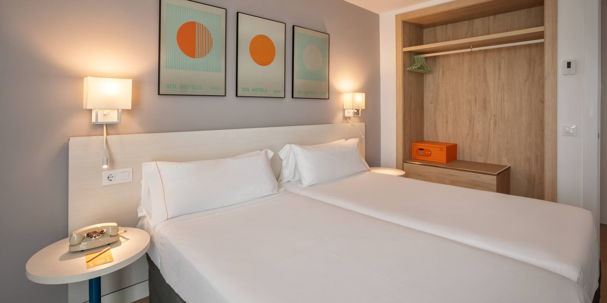 Bright hotel room with twin beds, modern art, open wardrobe, and safe.