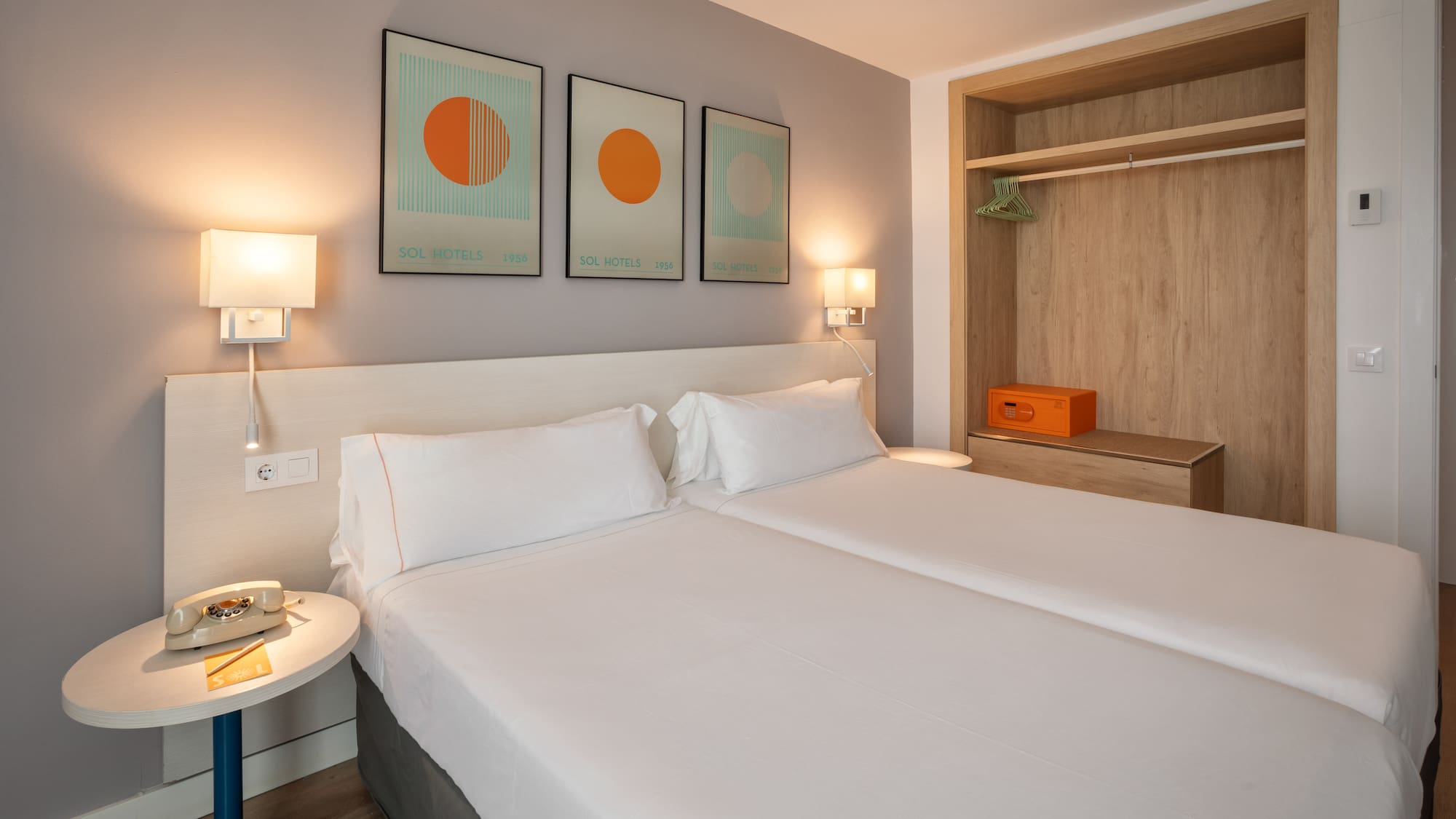 Bright hotel room with twin beds, modern art, open wardrobe, and safe.