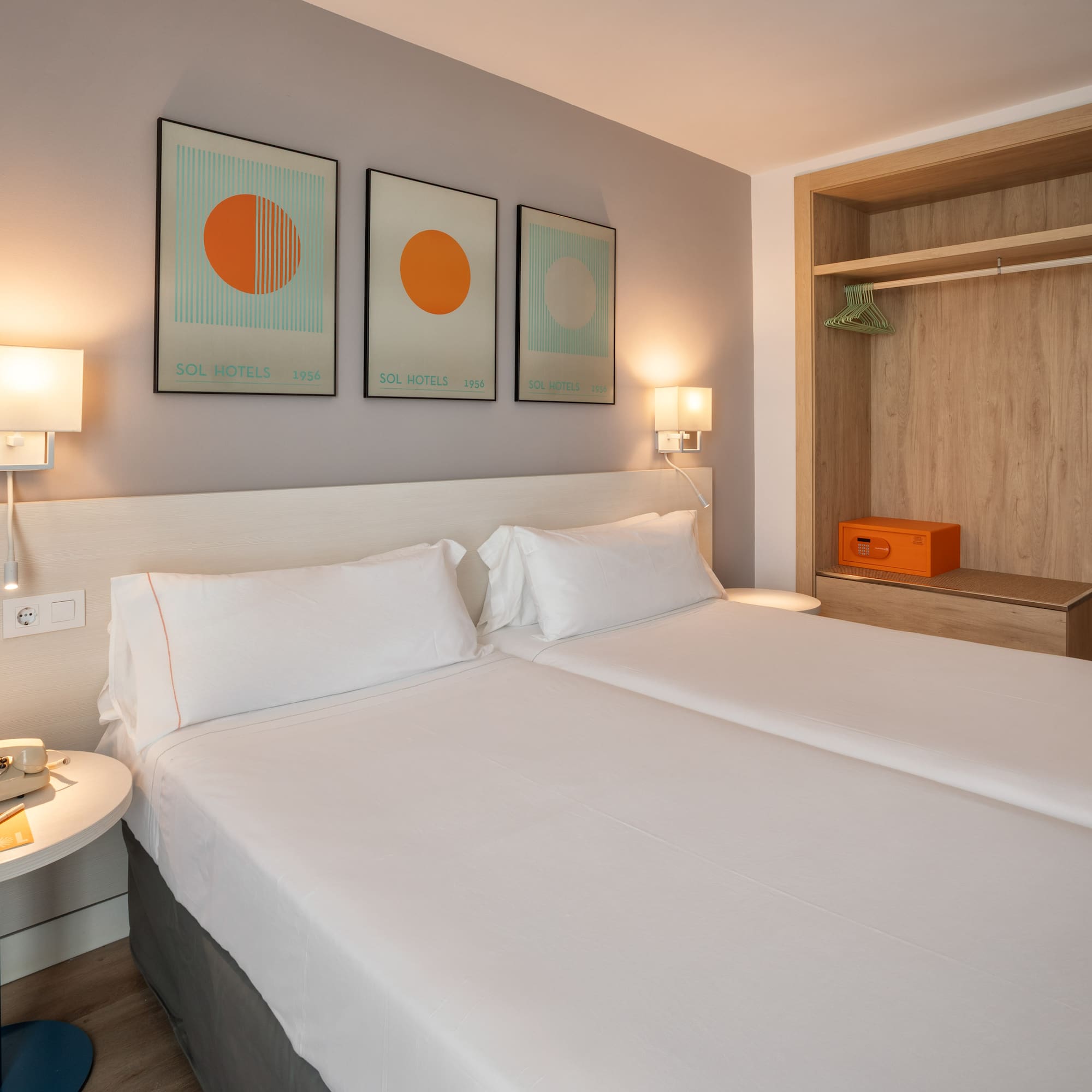 Bright hotel room with twin beds, modern art, open wardrobe, and safe.