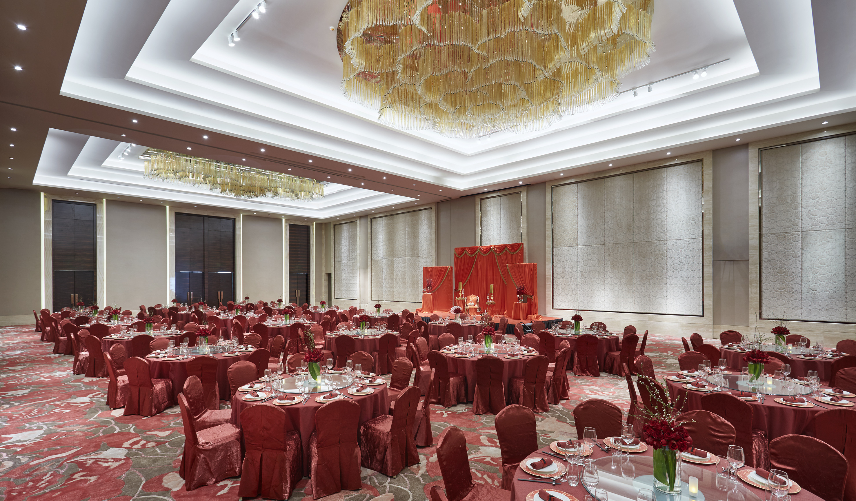 Grand ballroom with red tables, chairs, ornate chandelier, and event stage.