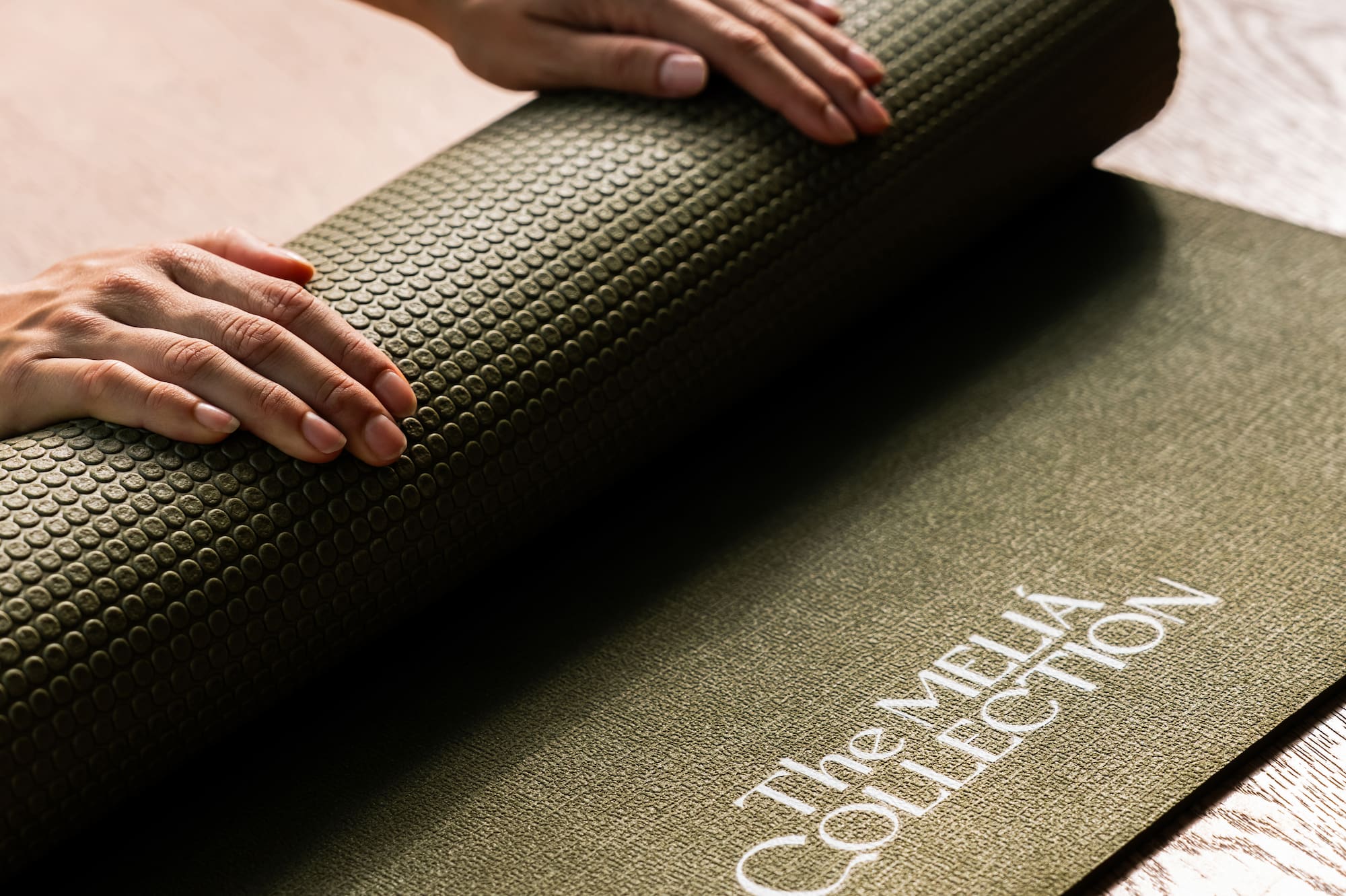 Hands rolling up textured olive green Meliá Collection yoga mat on wooden floor.