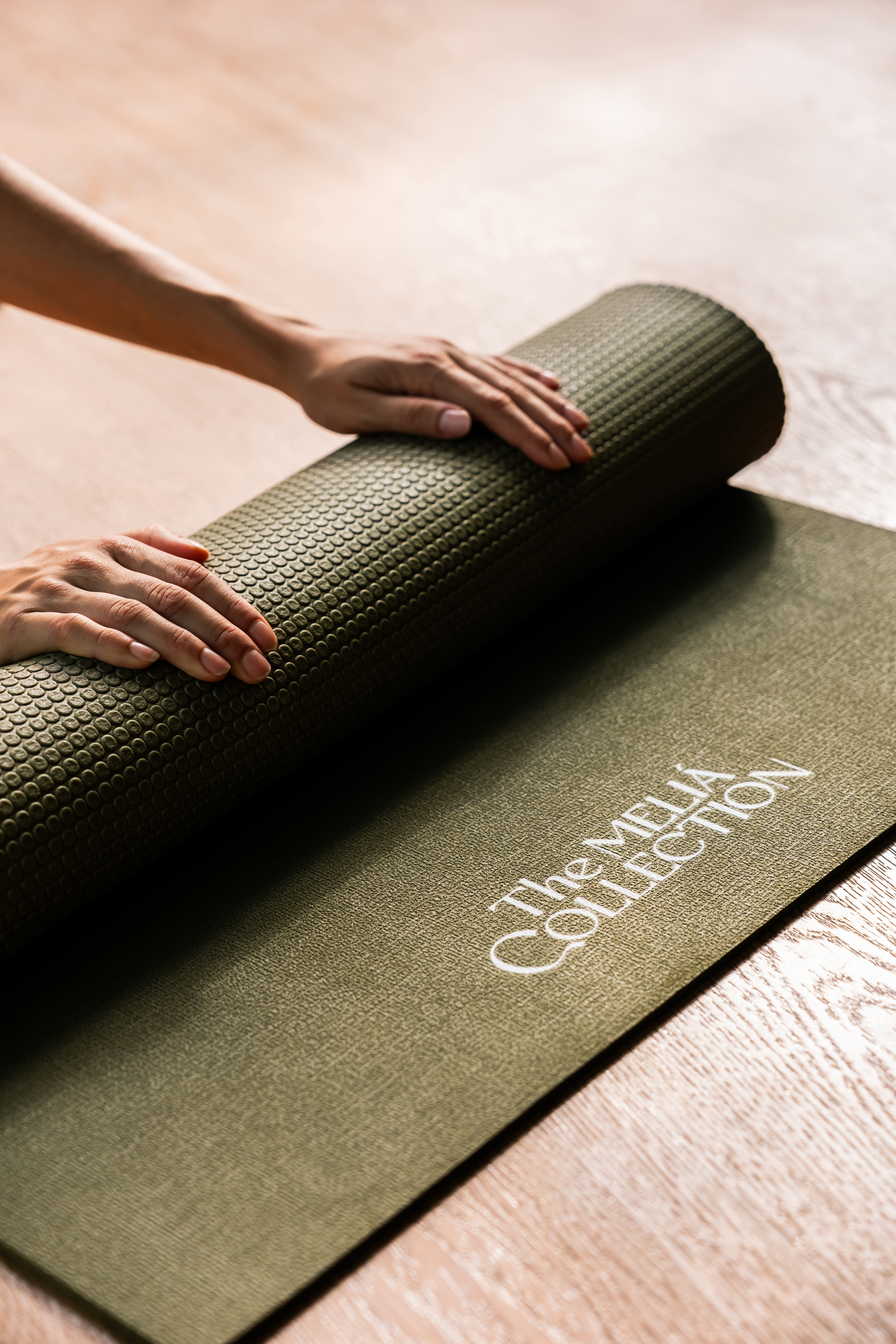 Hands rolling up textured olive green Meliá Collection yoga mat on wooden floor.