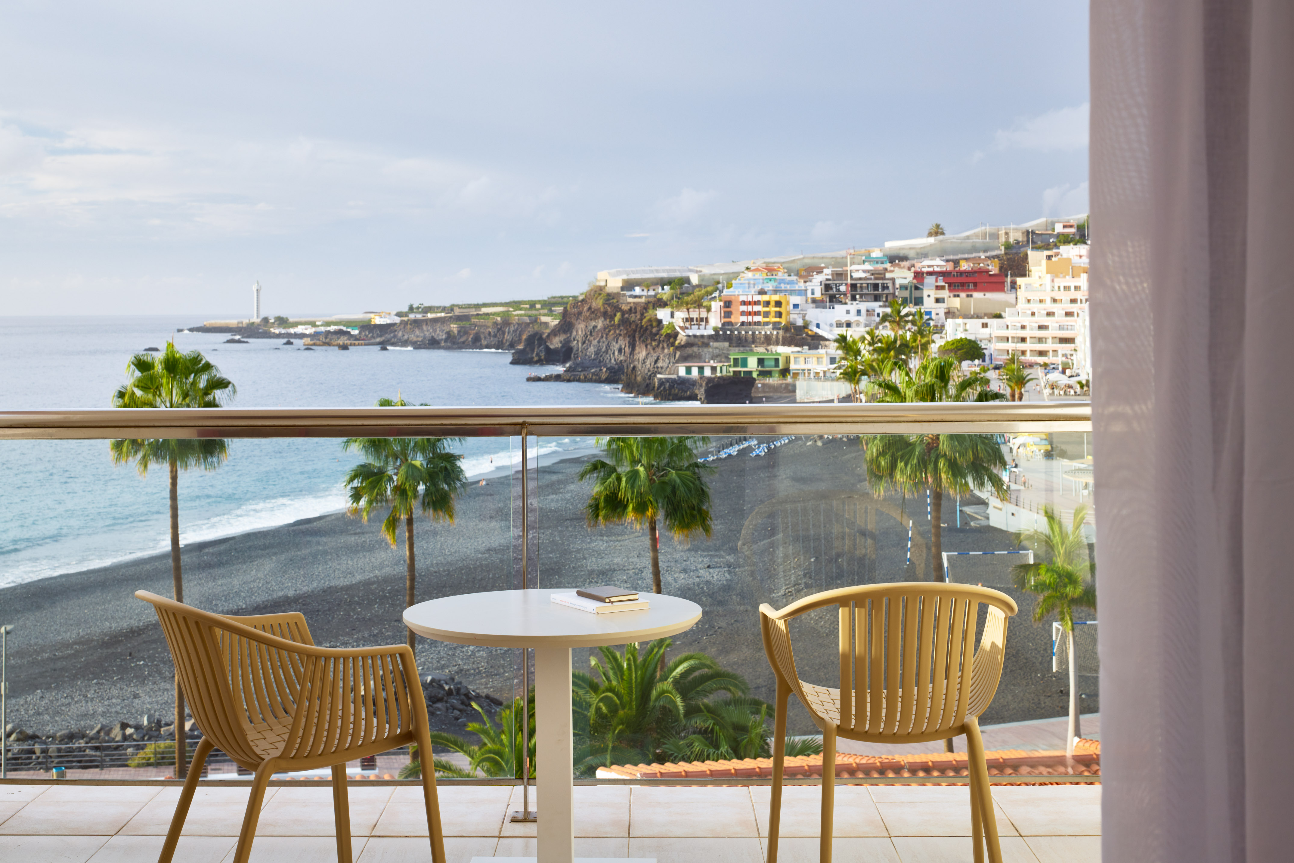 Hotel balcony ocean view: black sand beach, palm trees, and colorful coastal town.