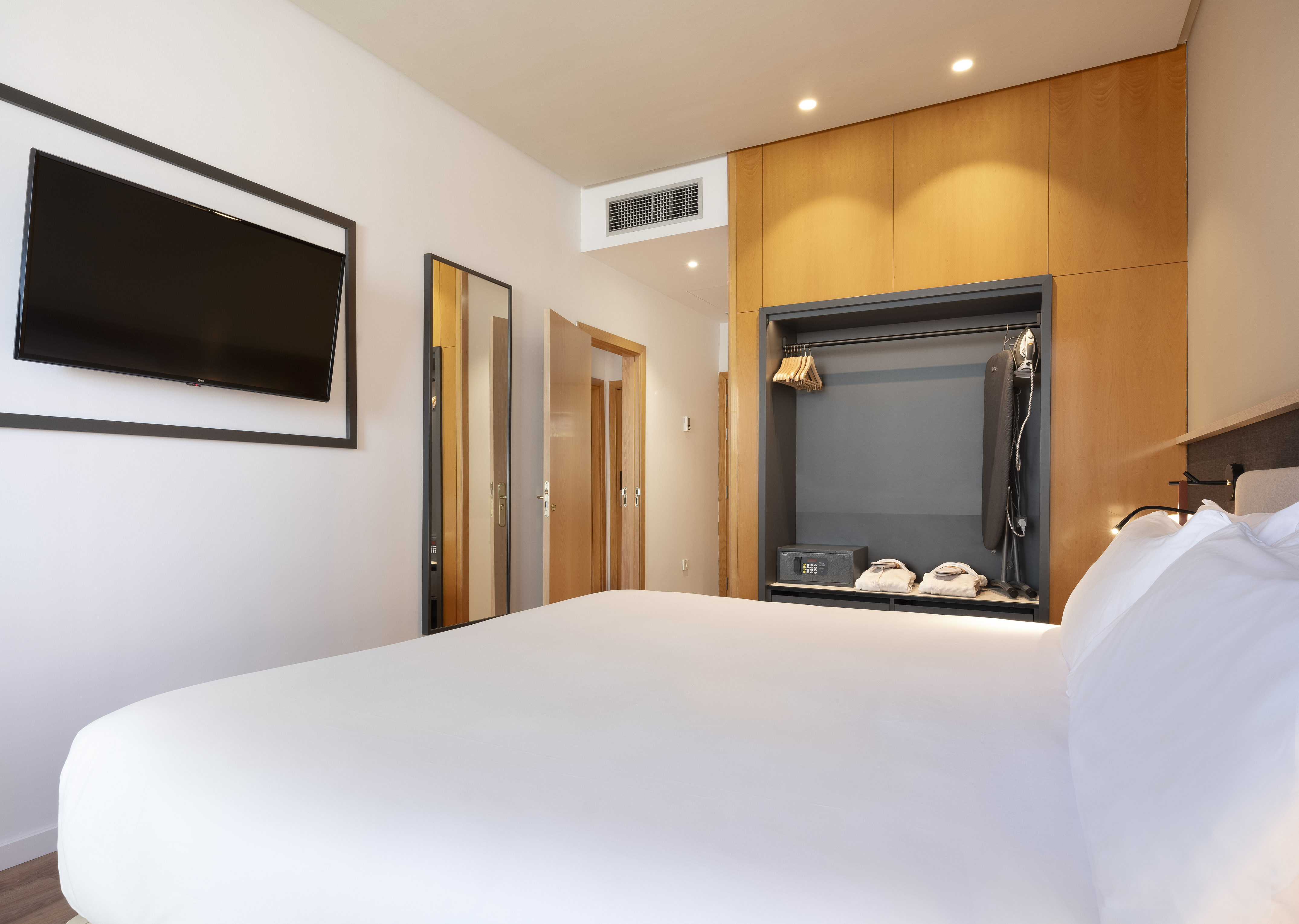 Spacious hotel room with white bed, wall TV, and open wardrobe with safe.