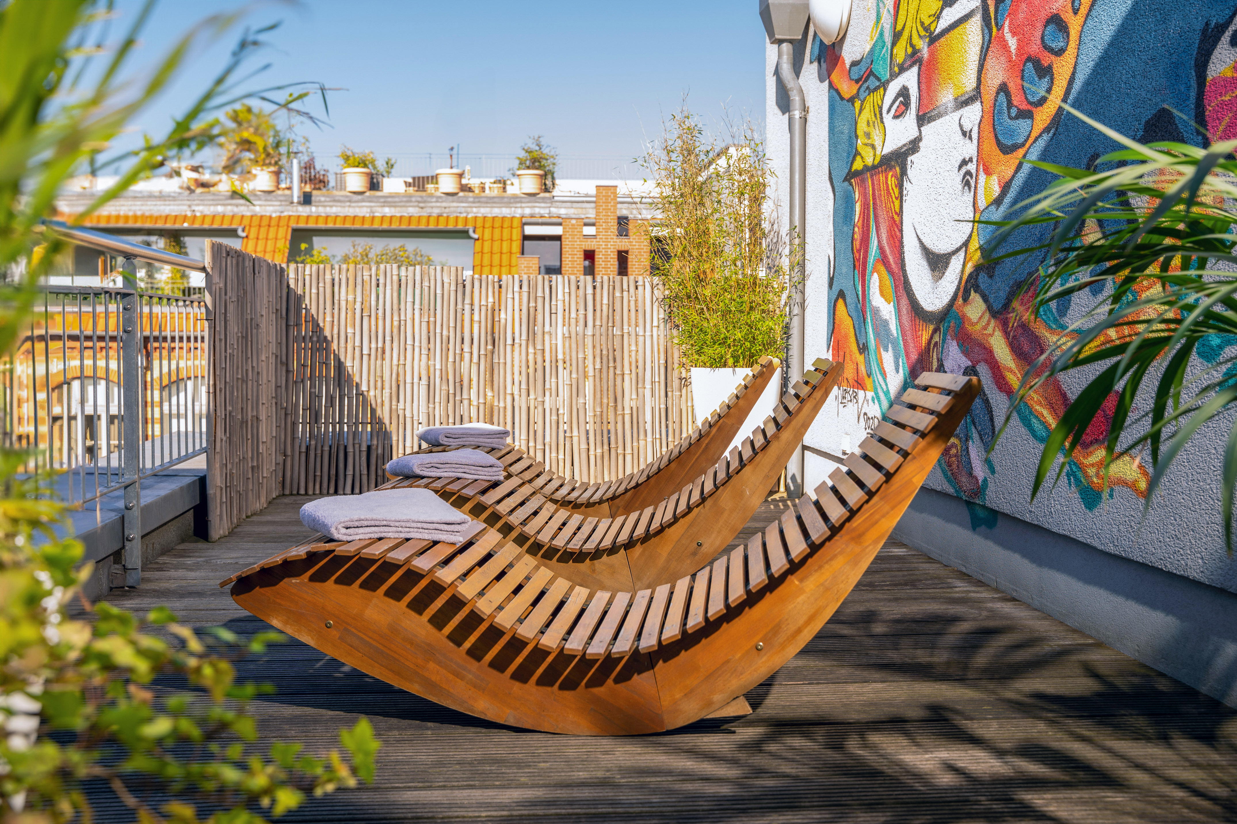 Sunny rooftop deck with wooden lounge chairs, bamboo fence, and colorful graffiti.
