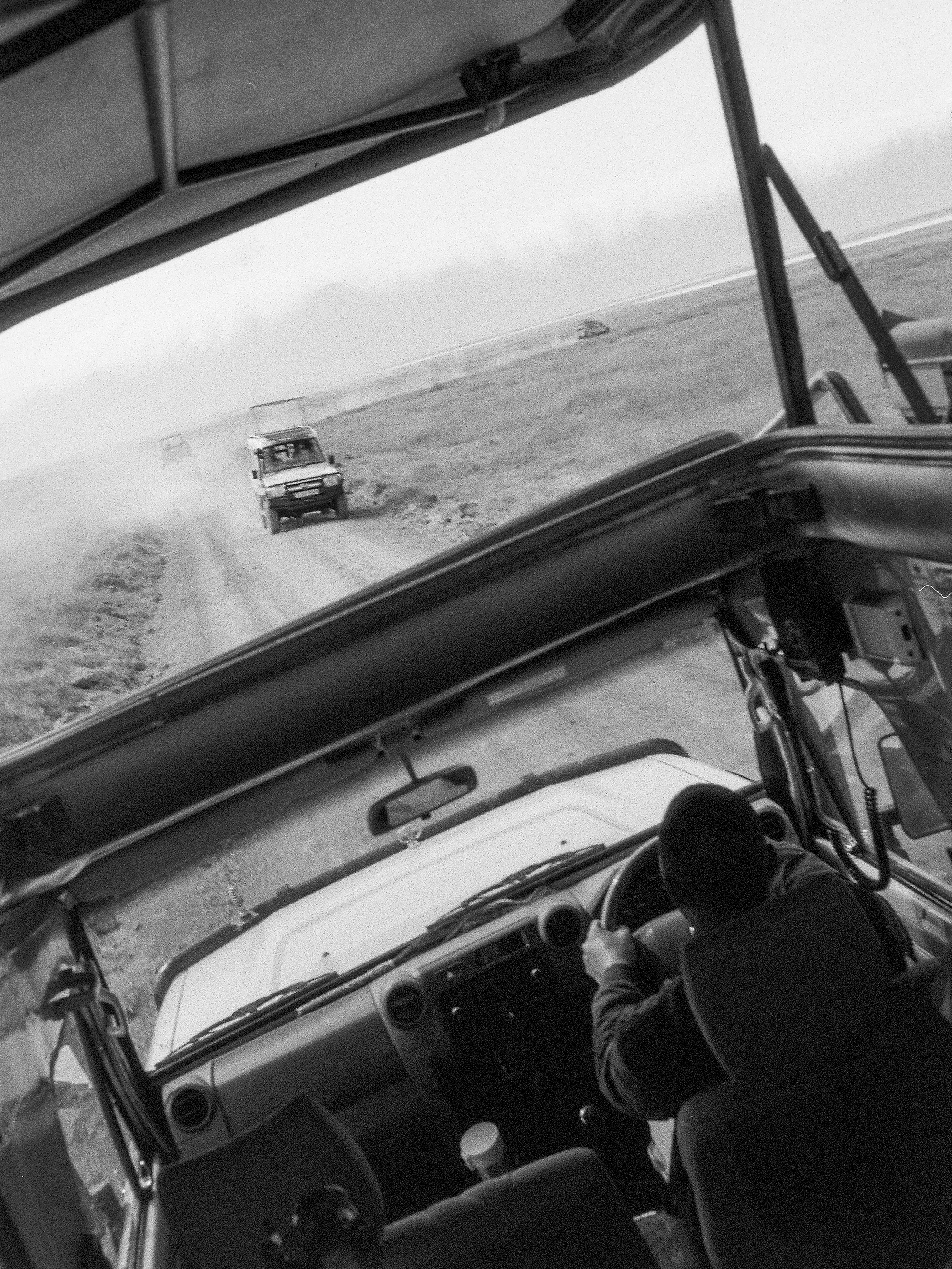 B&W safari vehicle, driver, following others on dusty off-road track.