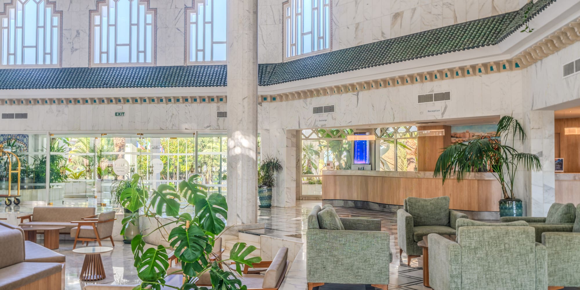 Bright hotel lobby with green armchairs, marble floor, and large arched windows.