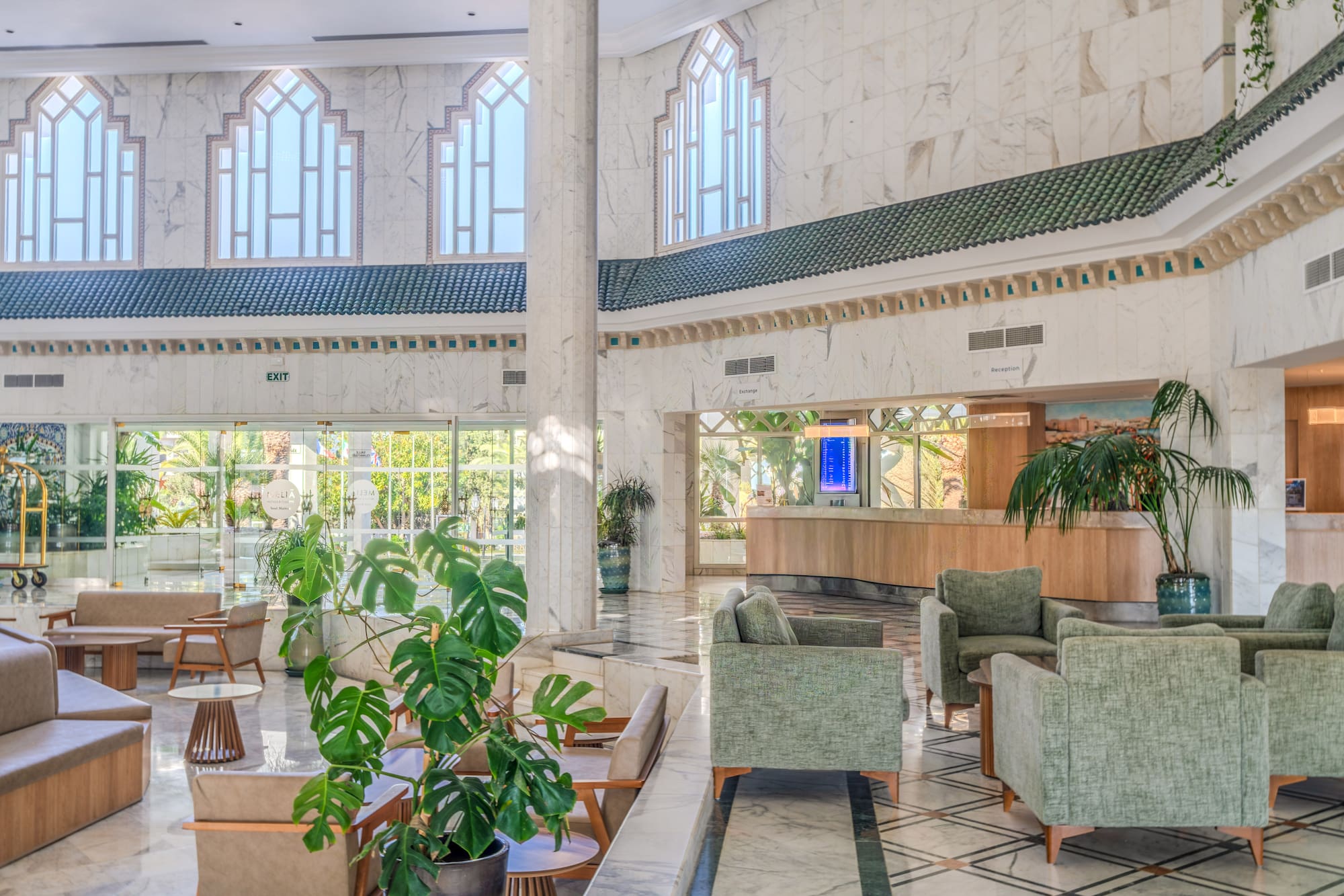 Bright hotel lobby with green armchairs, marble floor, and large arched windows.