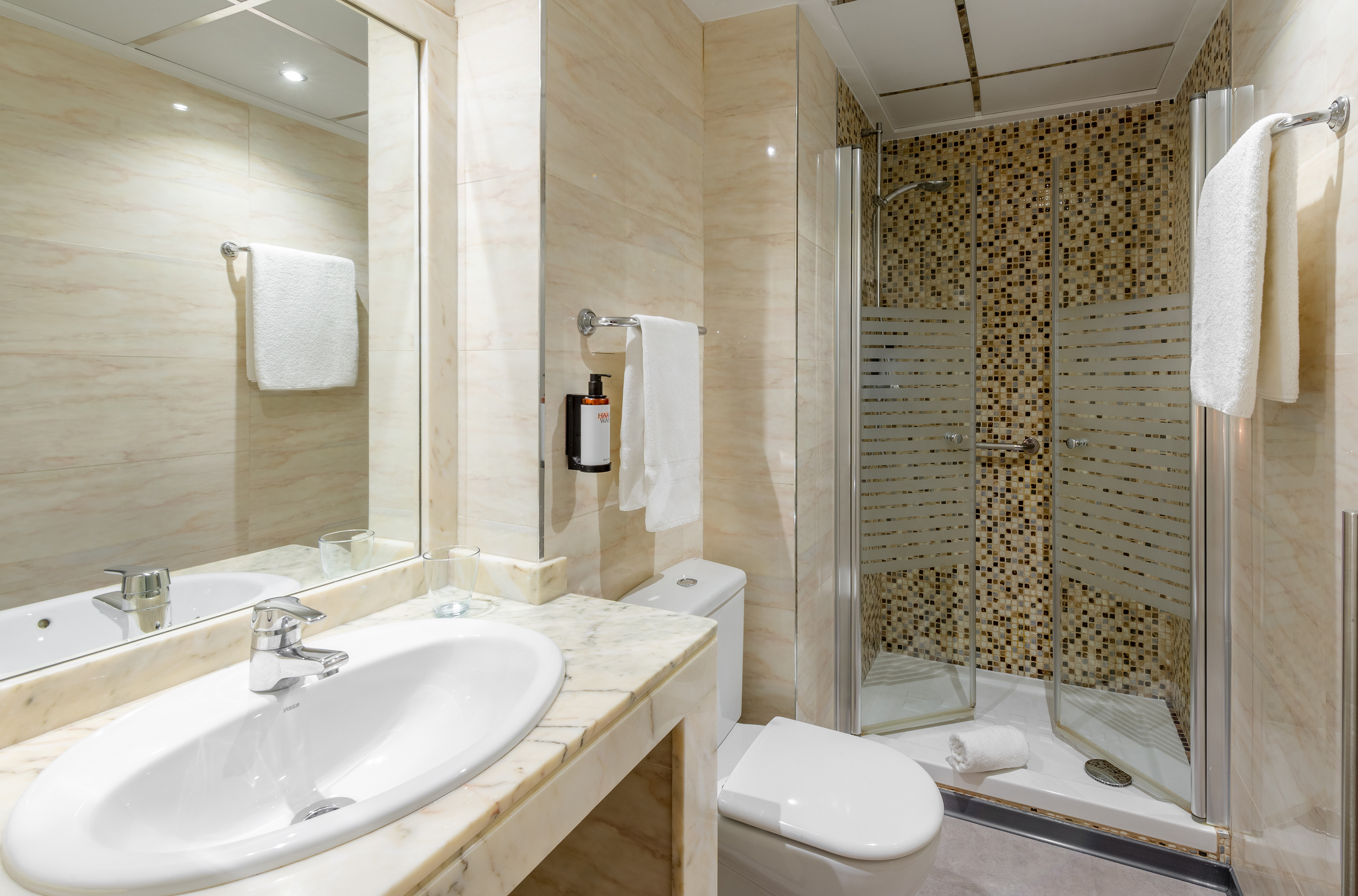 Modern hotel bathroom with marble vanity, sink, toilet, and mosaic tile shower.