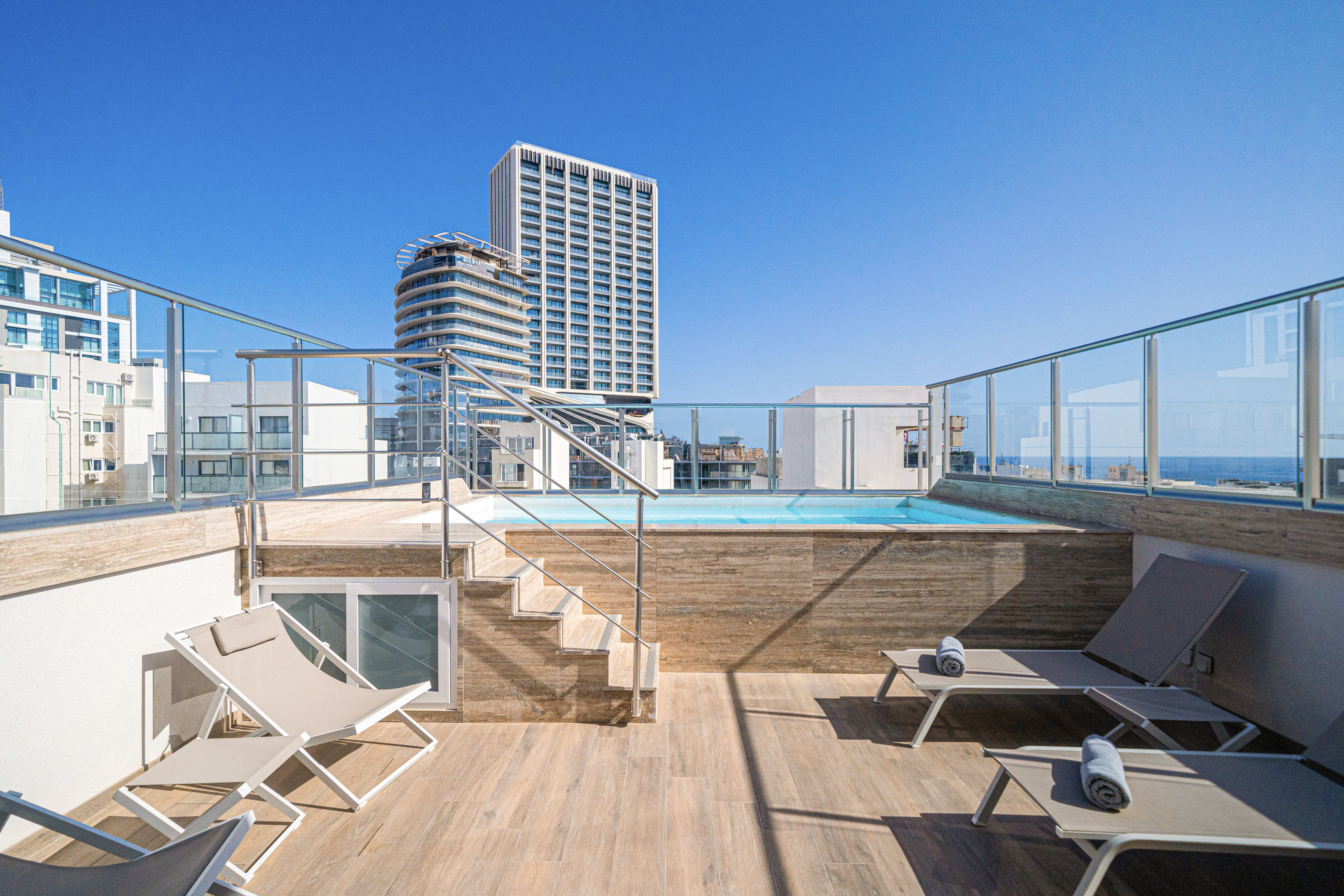 Rooftop pool terrace with sun loungers, city skyline, and sea views.