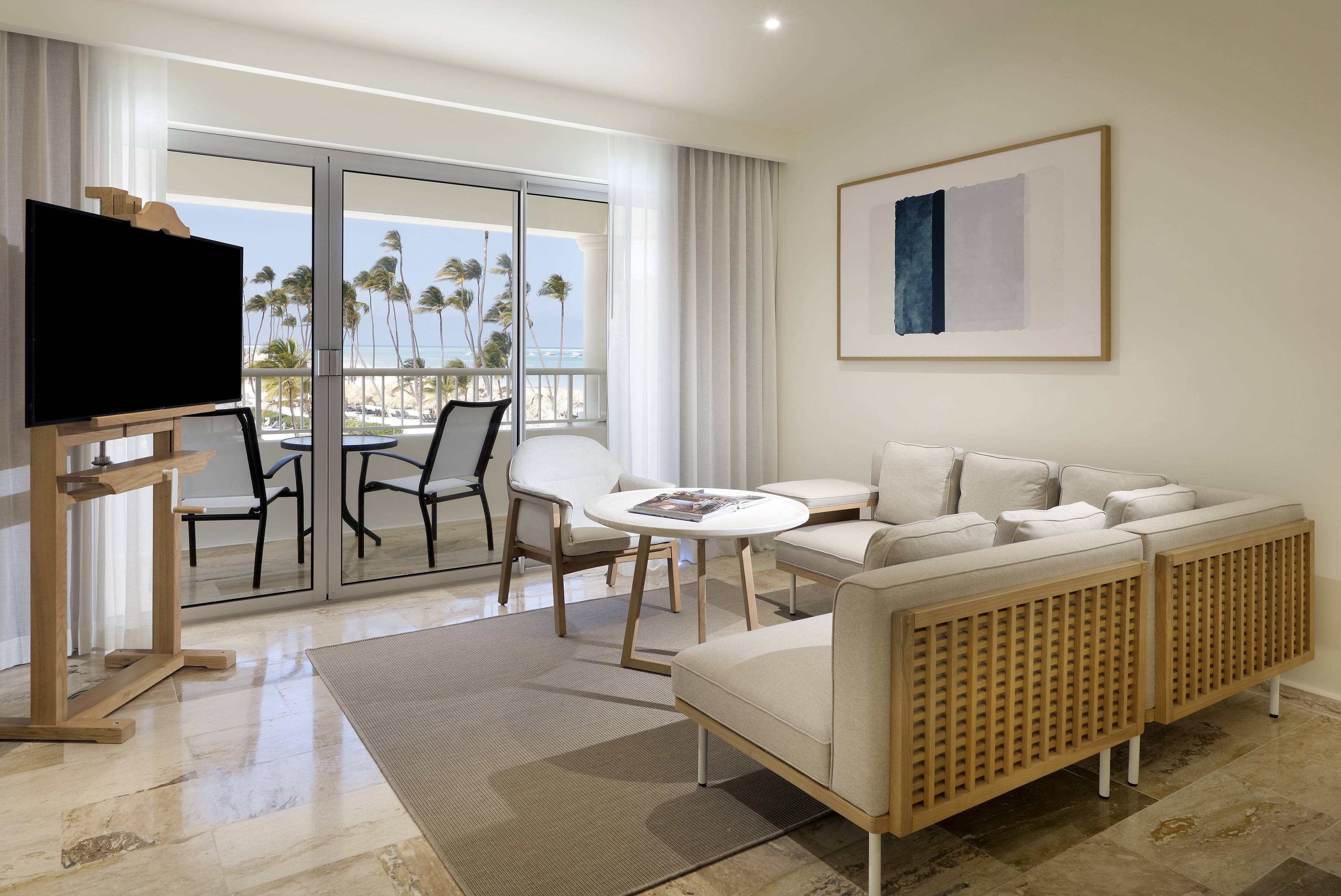 Modern hotel suite living room, ocean view balcony, palm trees, beach.
