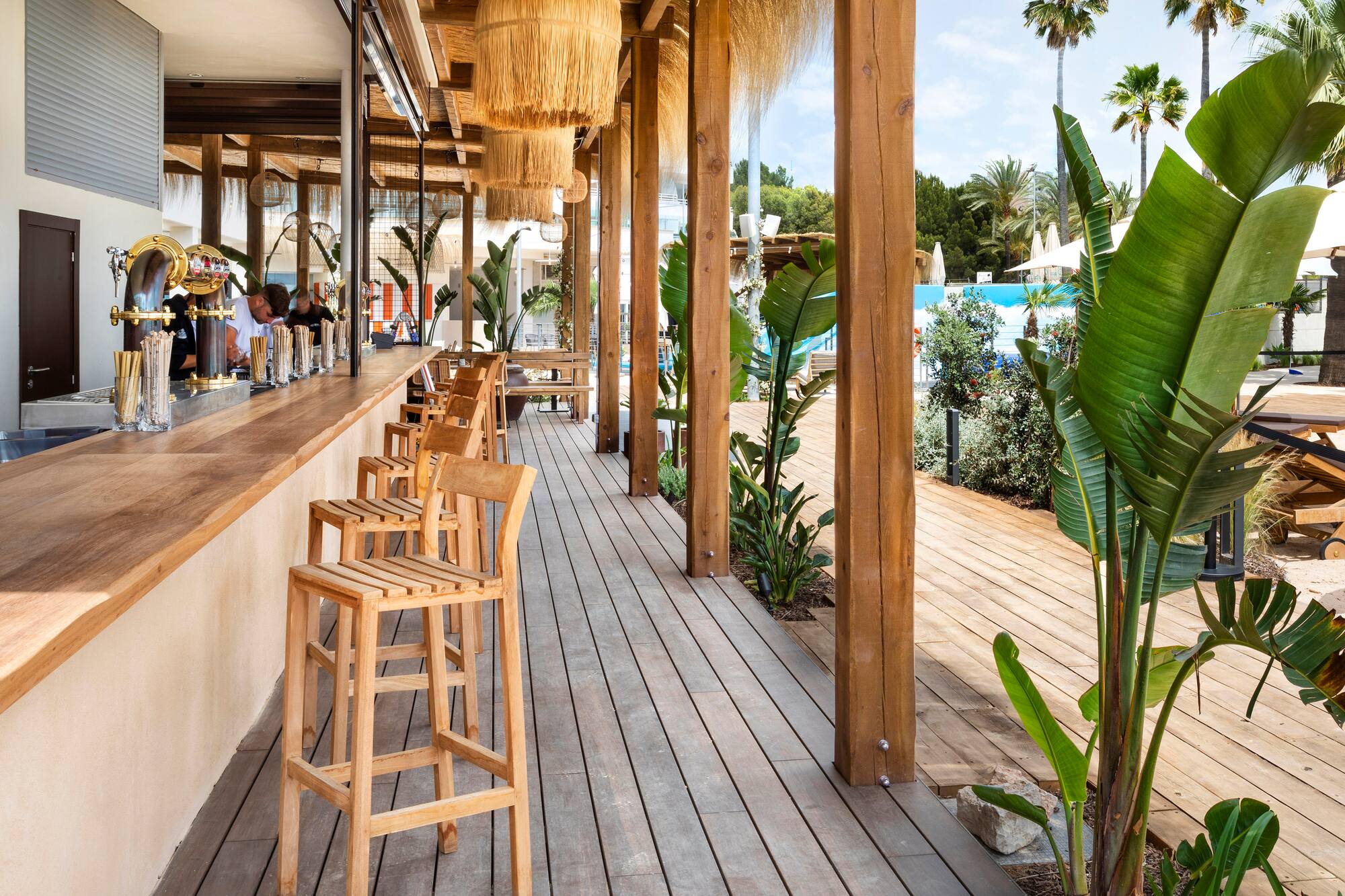 Sunny outdoor bar with wooden counter, deck, tropical plants, pool.