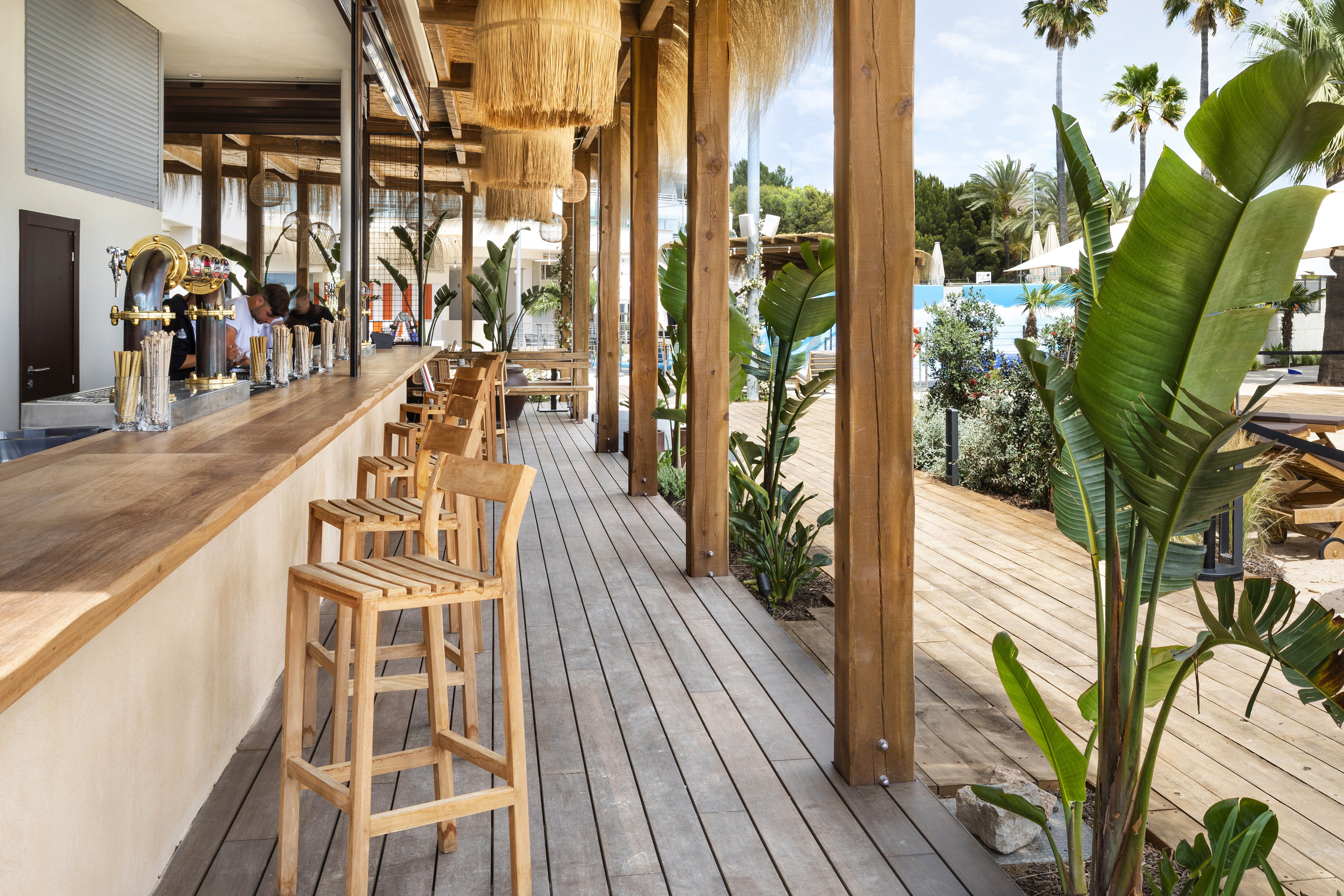 Sunny outdoor bar with wooden counter, deck, tropical plants, pool.