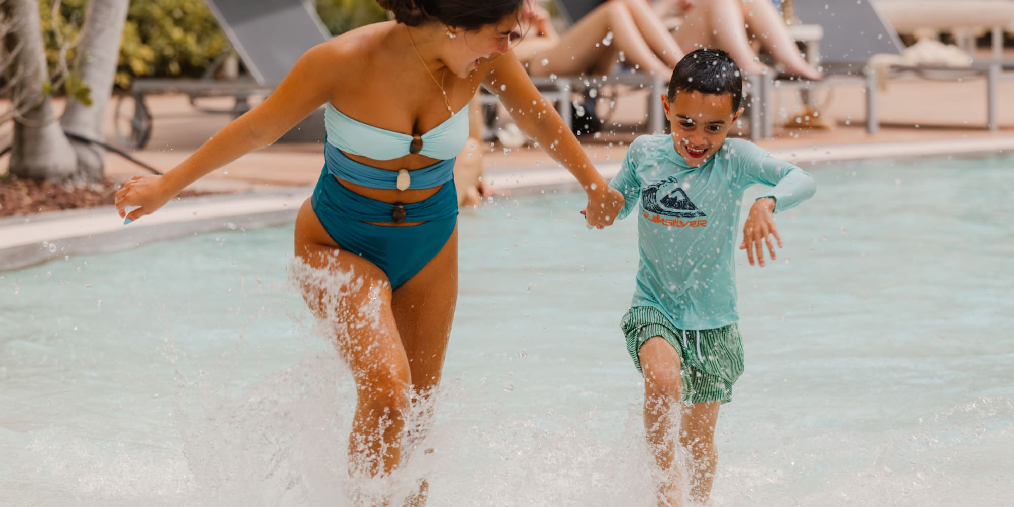 Happy woman and boy splashing in a resort pool, holding hands.
