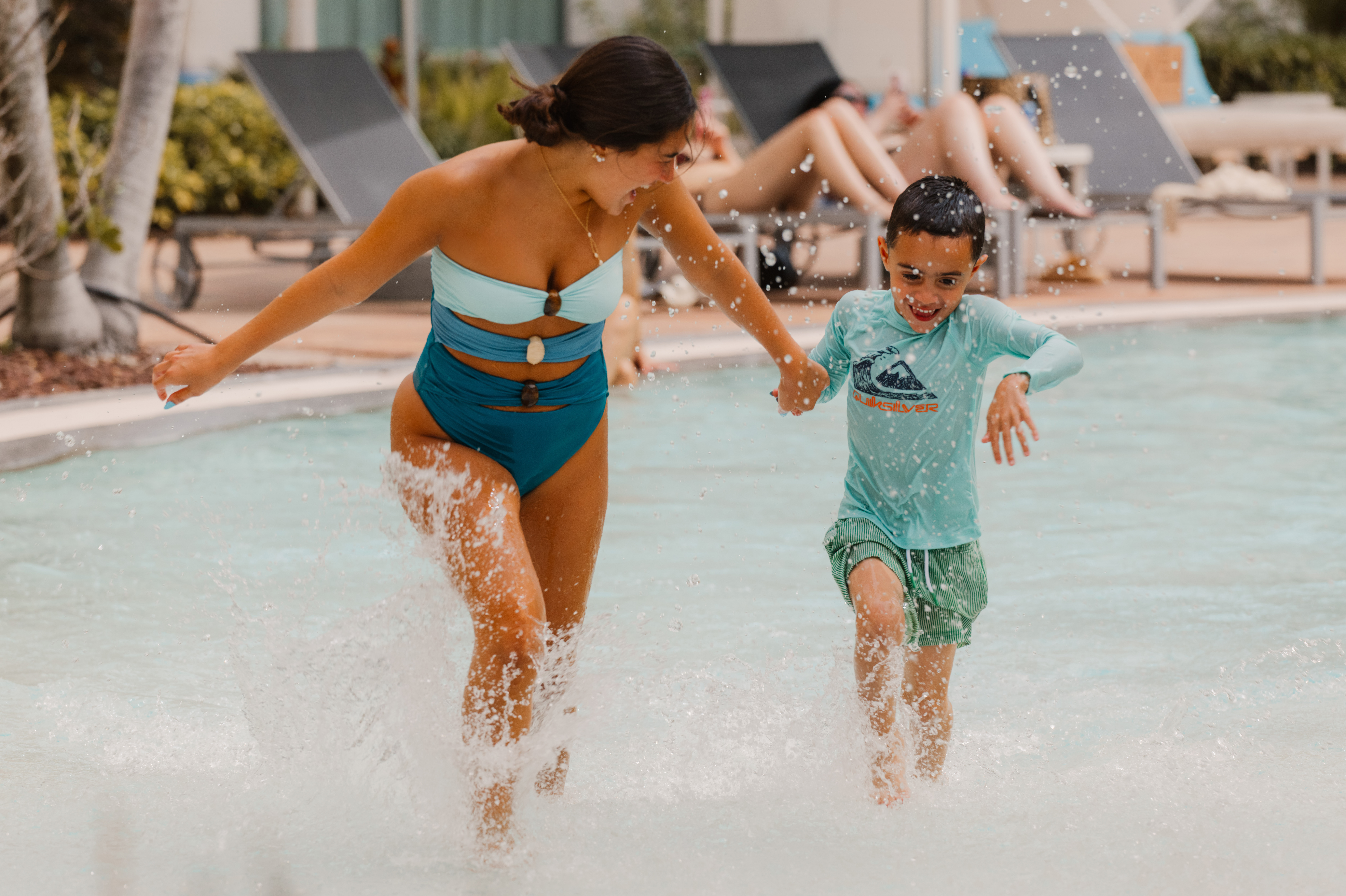 Happy woman and boy splashing in a resort pool, holding hands.