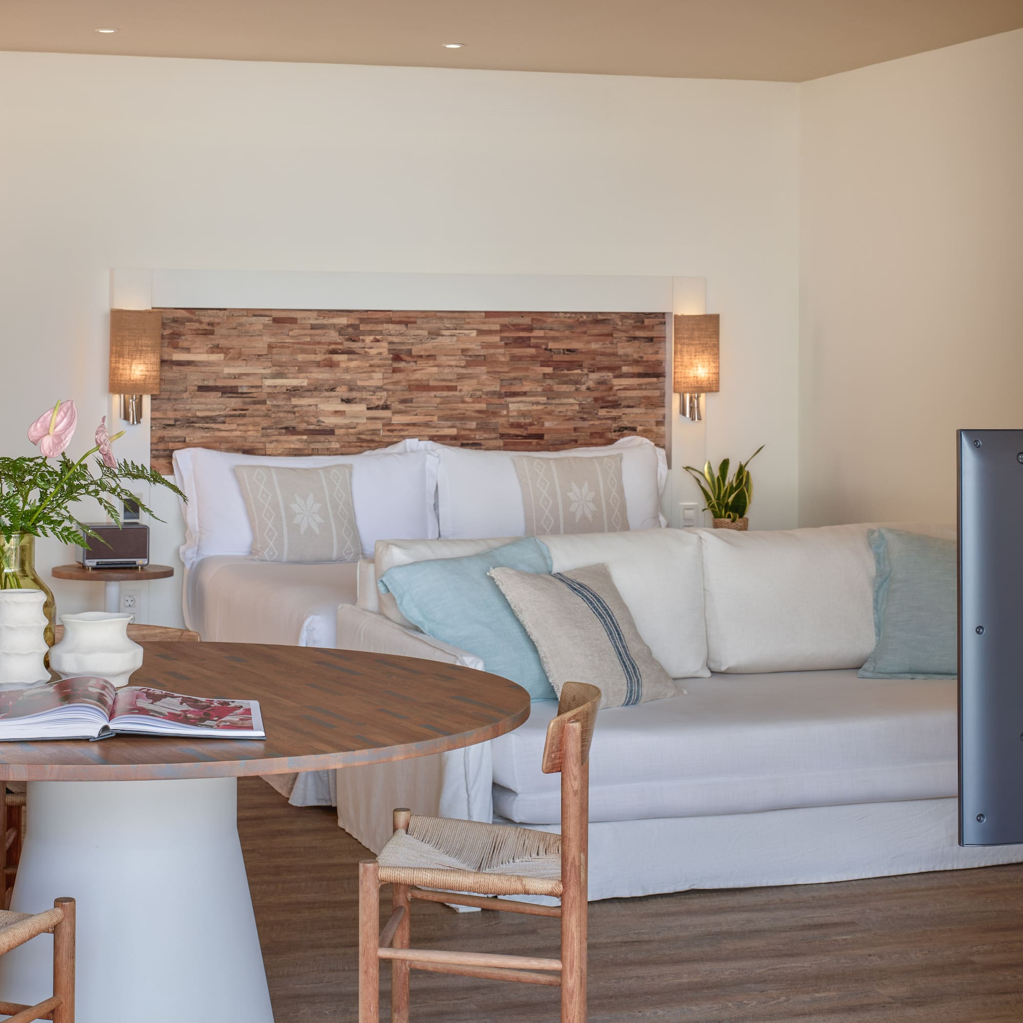 Bright hotel suite with wooden headboard, bed, white sofa, and dining table.