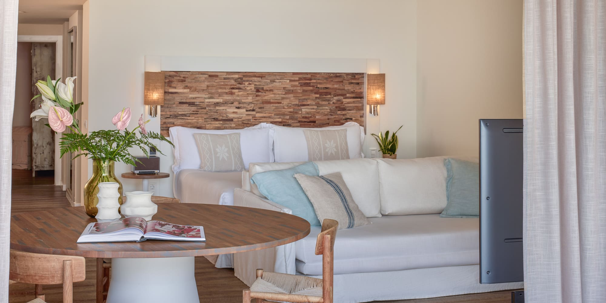 Bright hotel suite with wooden headboard, bed, white sofa, and dining table.