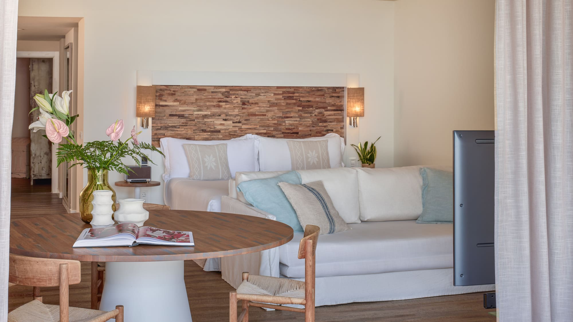 Bright hotel suite with wooden headboard, bed, white sofa, and dining table.
