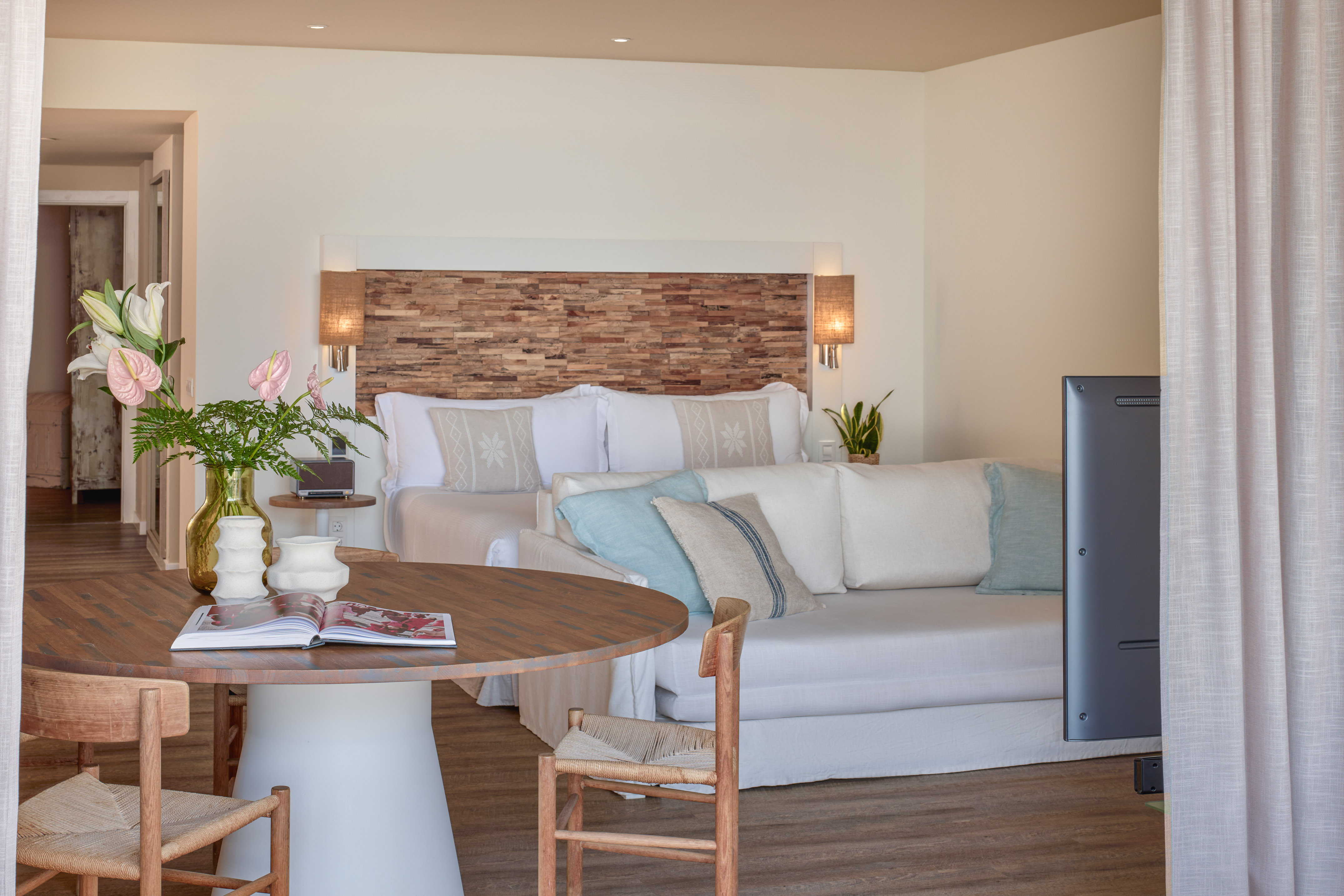 Bright hotel suite with wooden headboard, bed, white sofa, and dining table.
