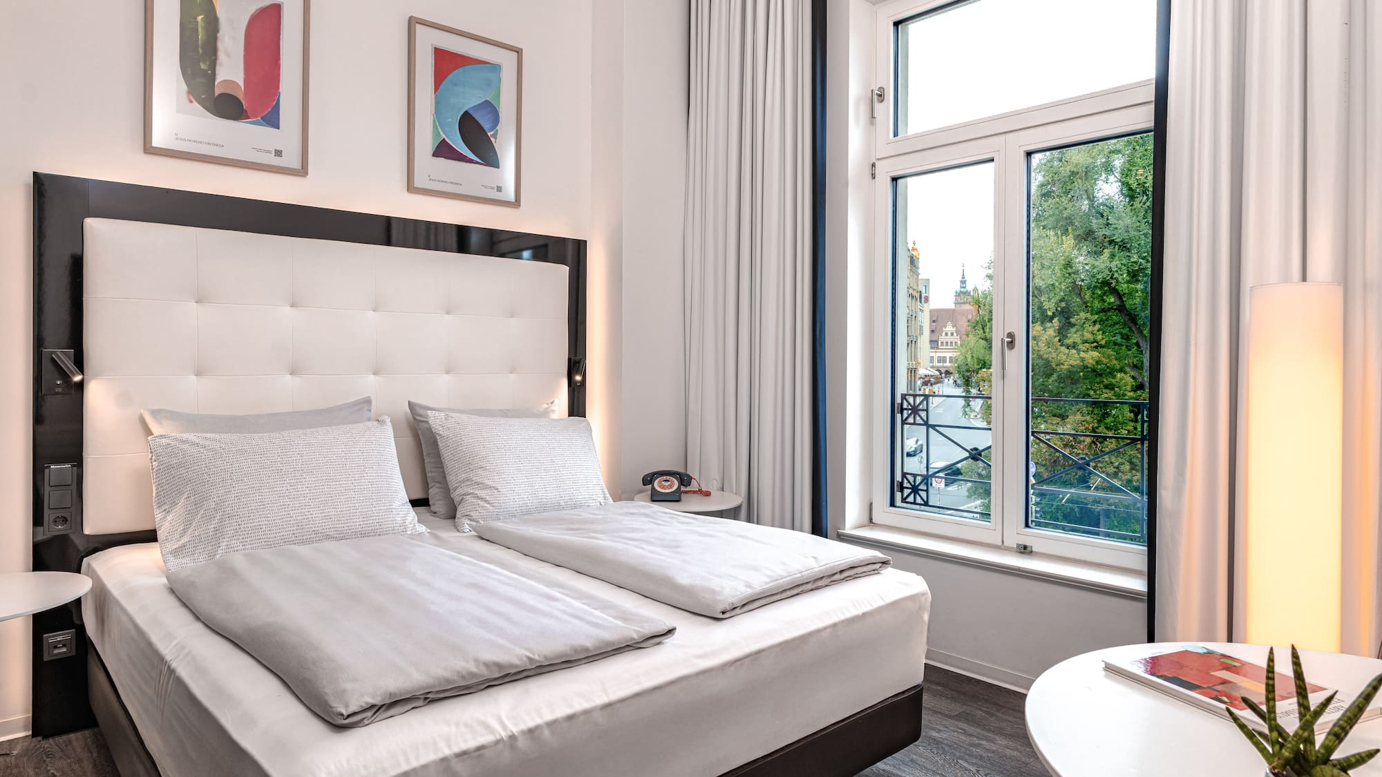 Modern hotel room with king bed, white bedding, artwork, and window city view.