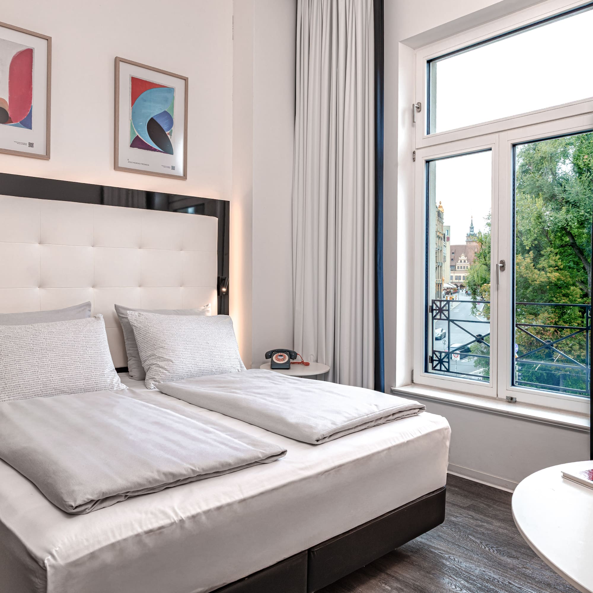 Modern hotel room with king bed, white bedding, artwork, and window city view.