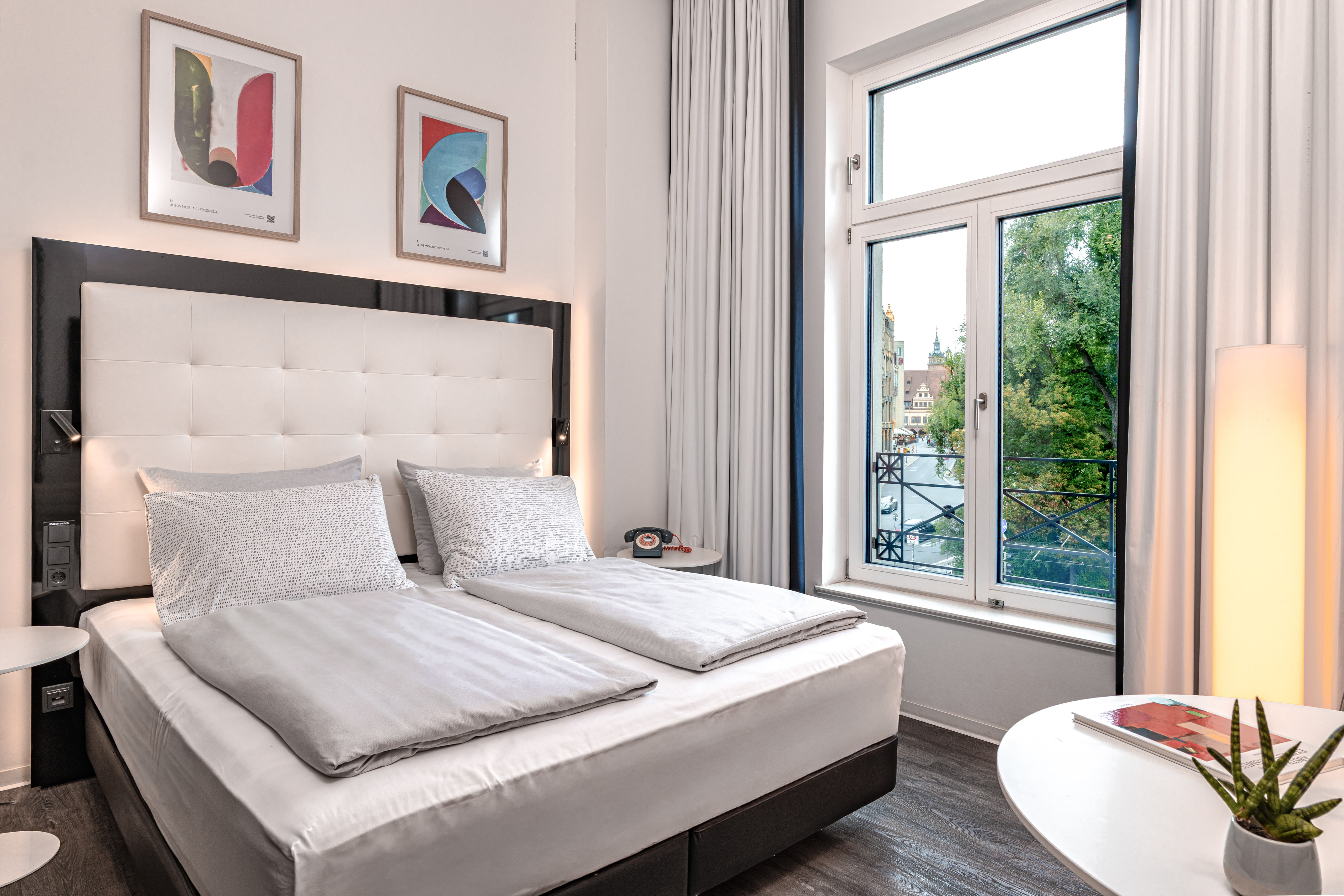 Modern hotel room with large bed, city view window, and contemporary decor.
