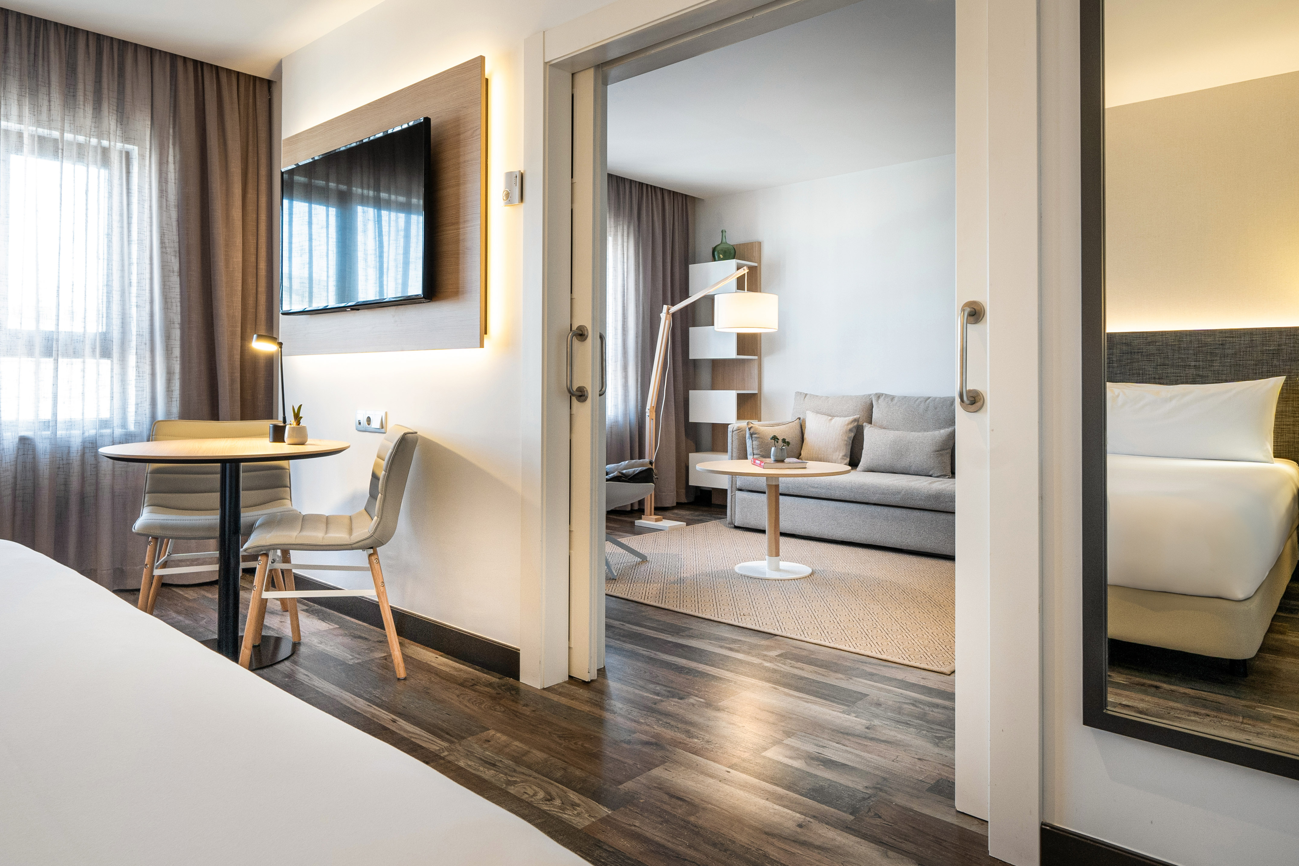 Modern hotel suite with bedroom, TV, and connected living room with sofa.