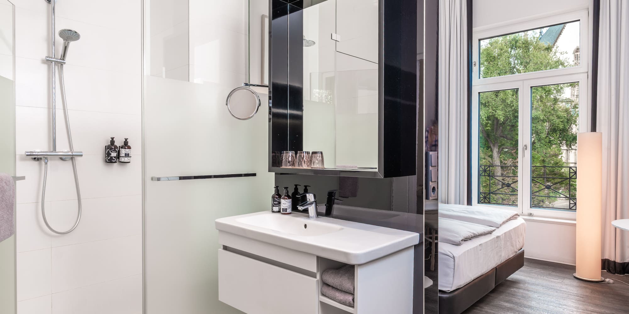 Modern hotel room with walk-in shower, vanity sink, and bed by a large window.