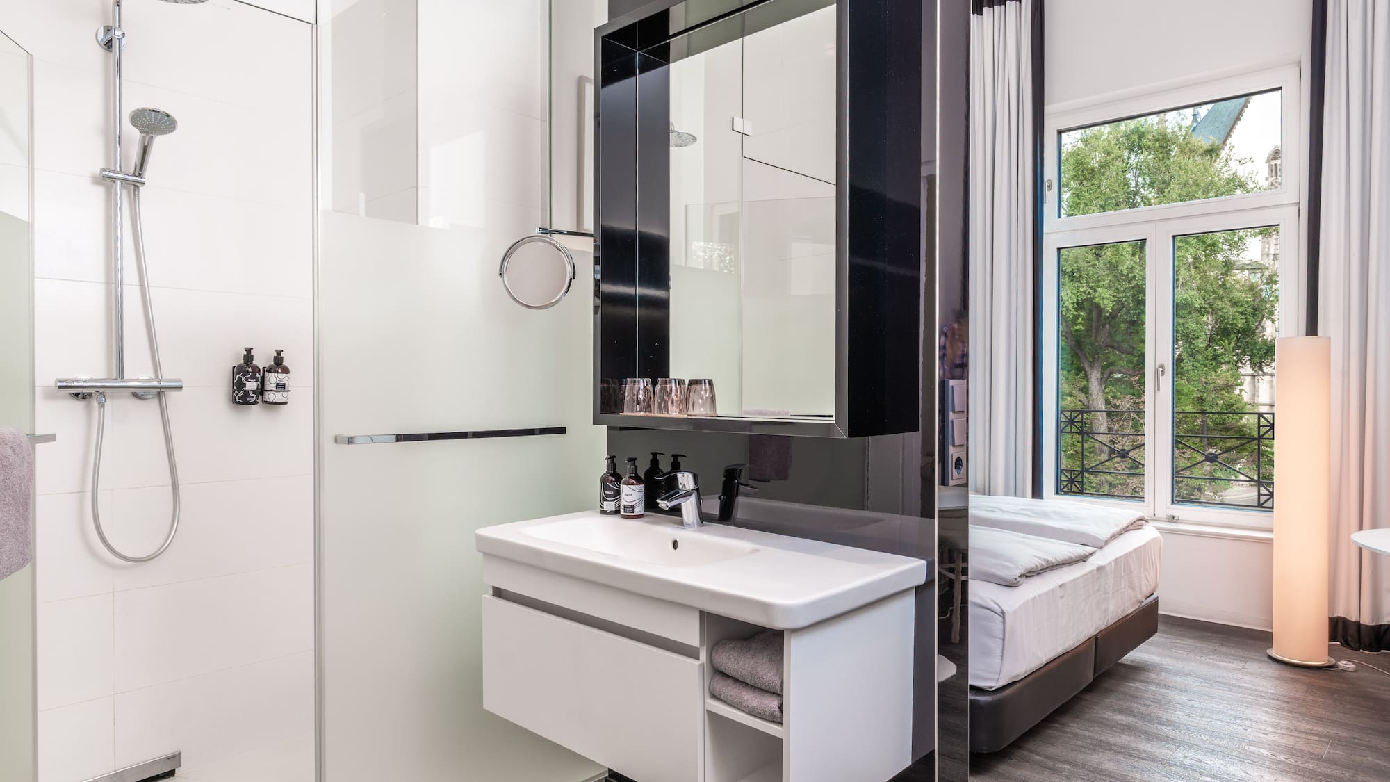 Modern hotel room: shower, vanity, and bed with window view.