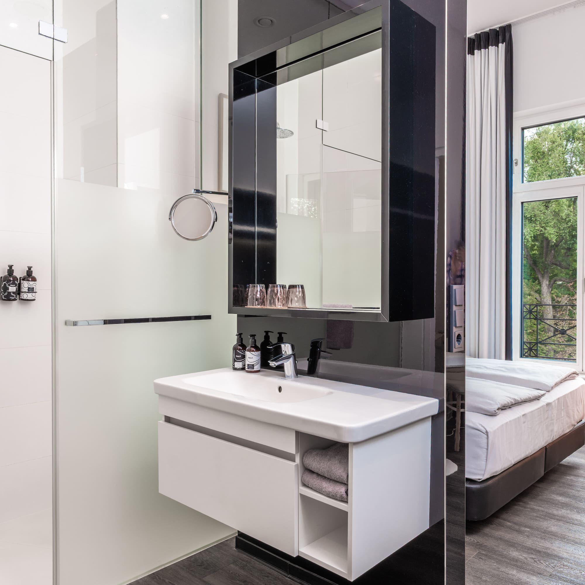 Modern hotel room: shower, vanity, and bed with window view.
