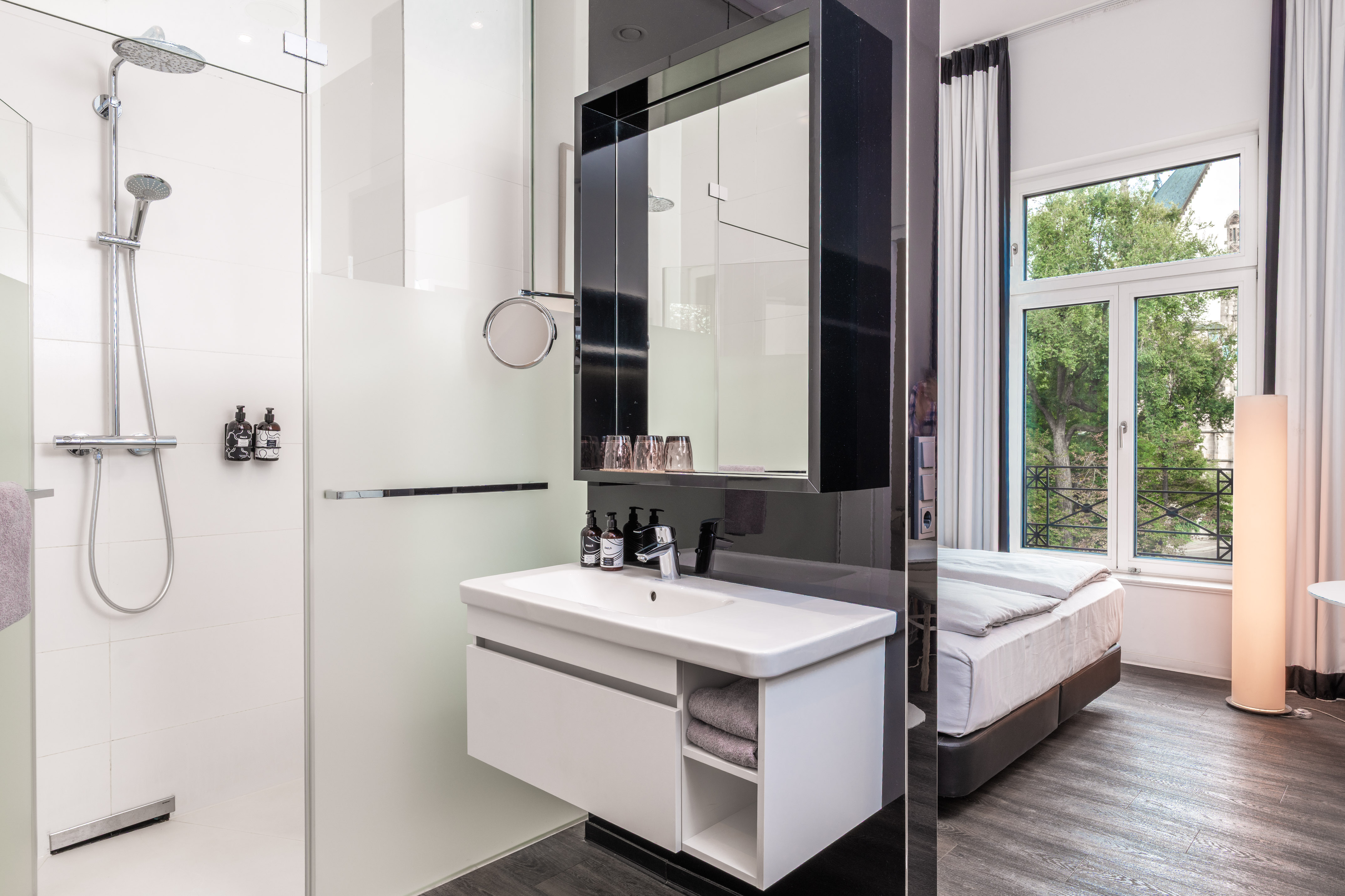 Modern hotel room with walk-in shower, vanity sink, and bed by a large window.