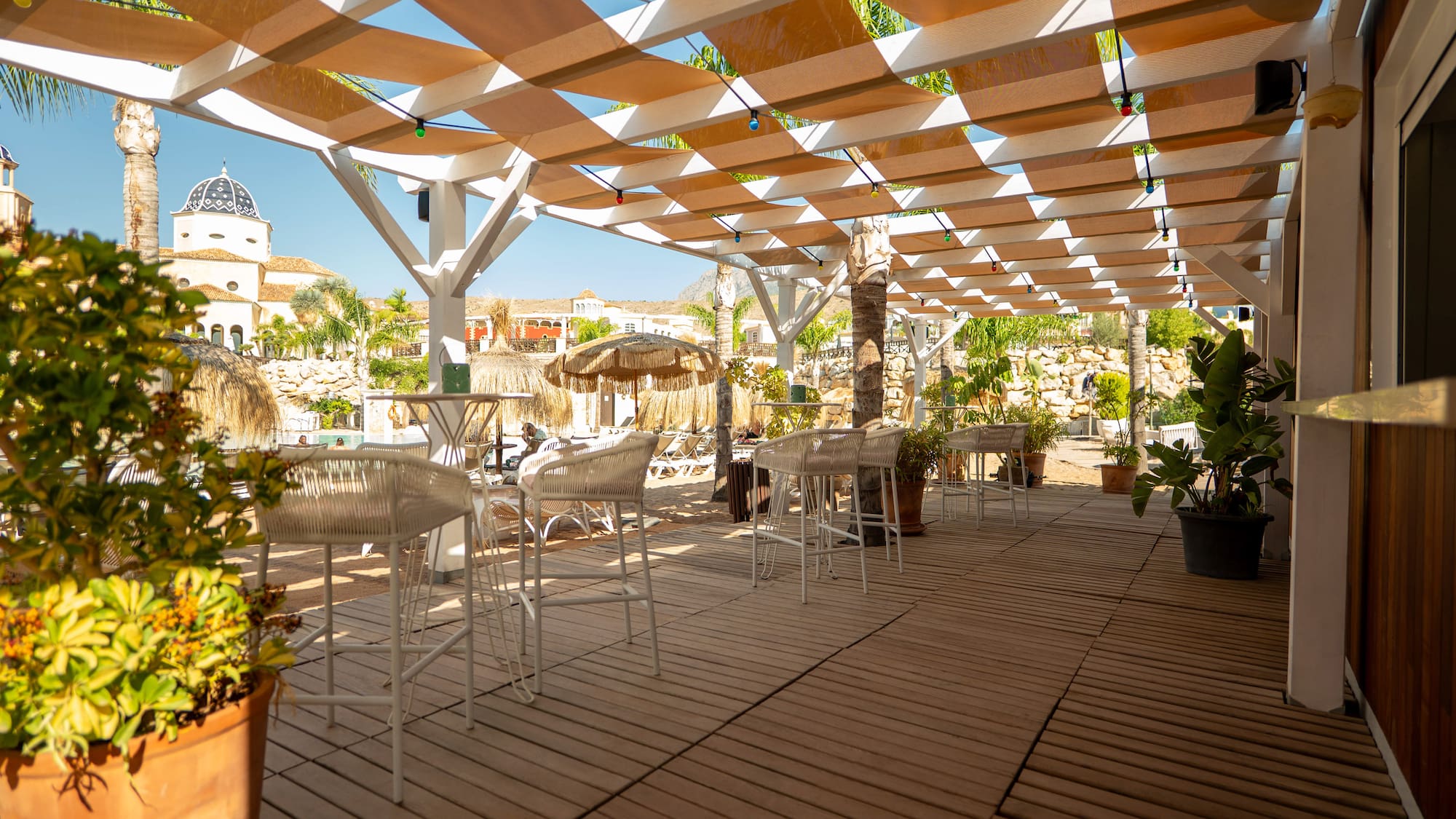 Resort outdoor bar with shaded pergola, wooden deck, white bar stools, and pool area.