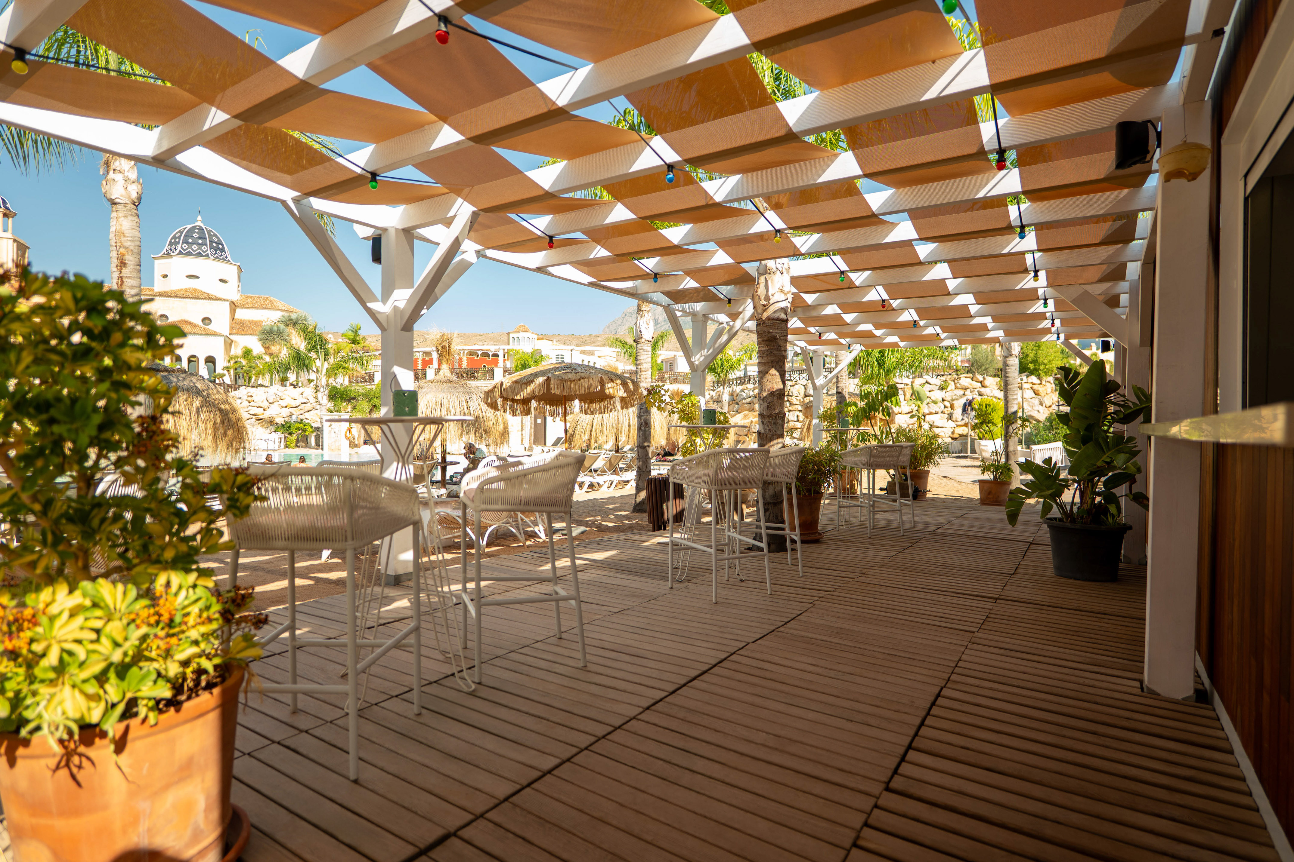 Resort outdoor bar with shaded pergola, wooden deck, white bar stools, and pool area.