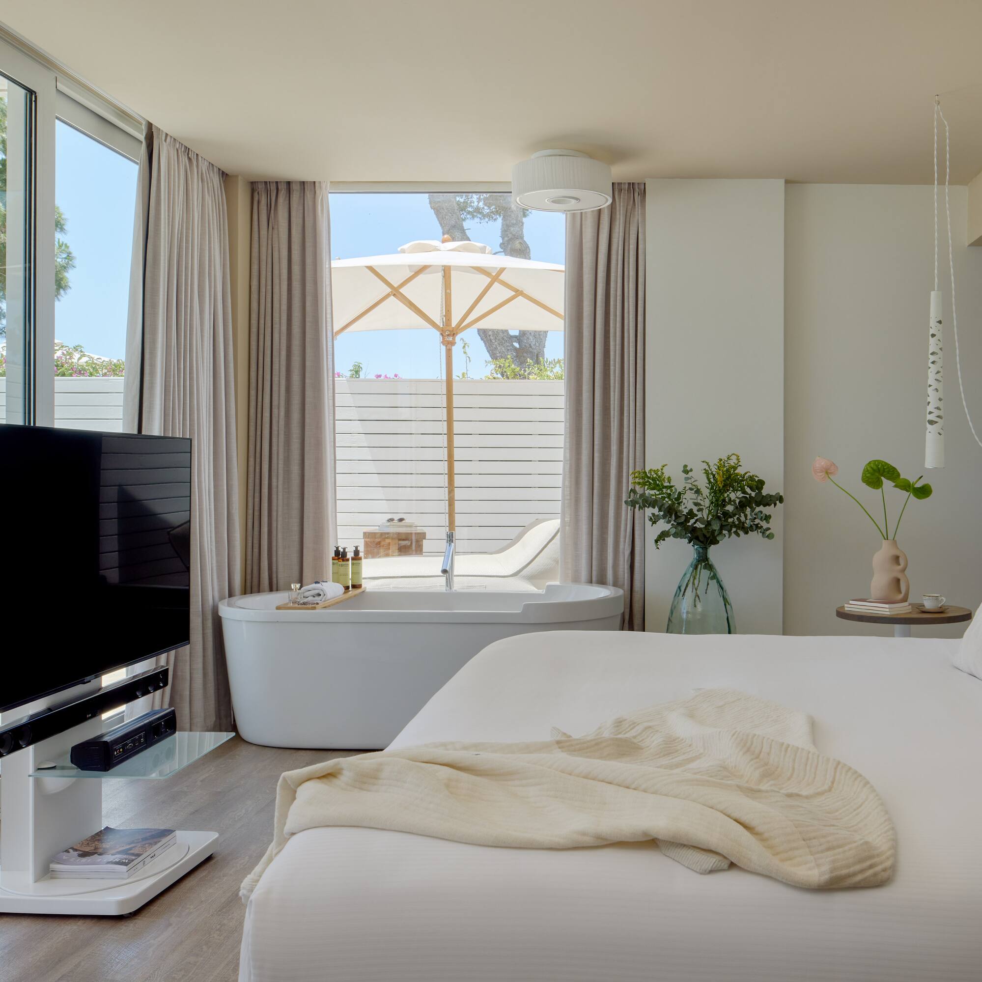 Modern hotel suite with freestanding tub, private patio, white decor, and TV.