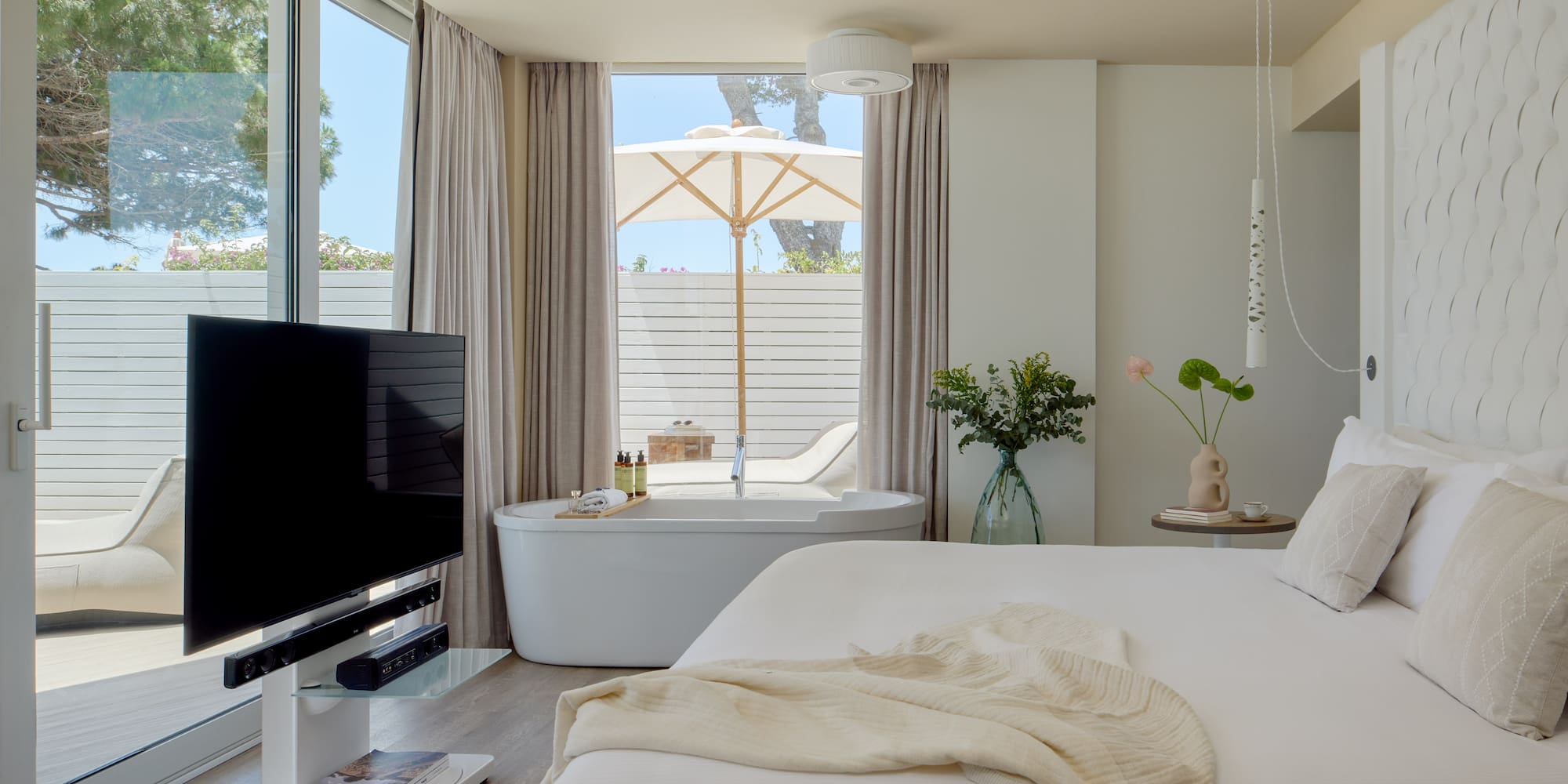 Modern hotel suite with freestanding tub, private patio, white decor, and TV.