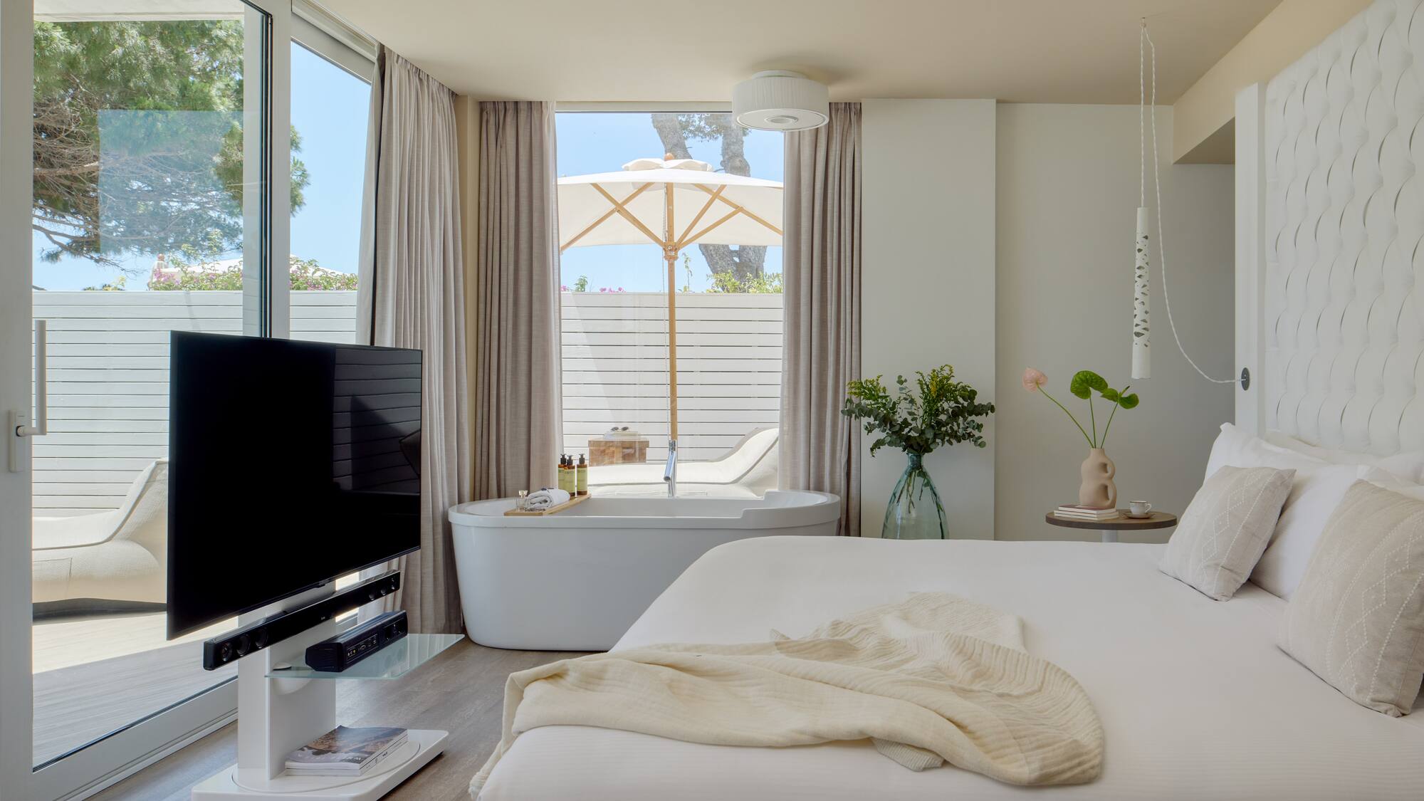 Modern hotel suite with freestanding tub, private patio, white decor, and TV.
