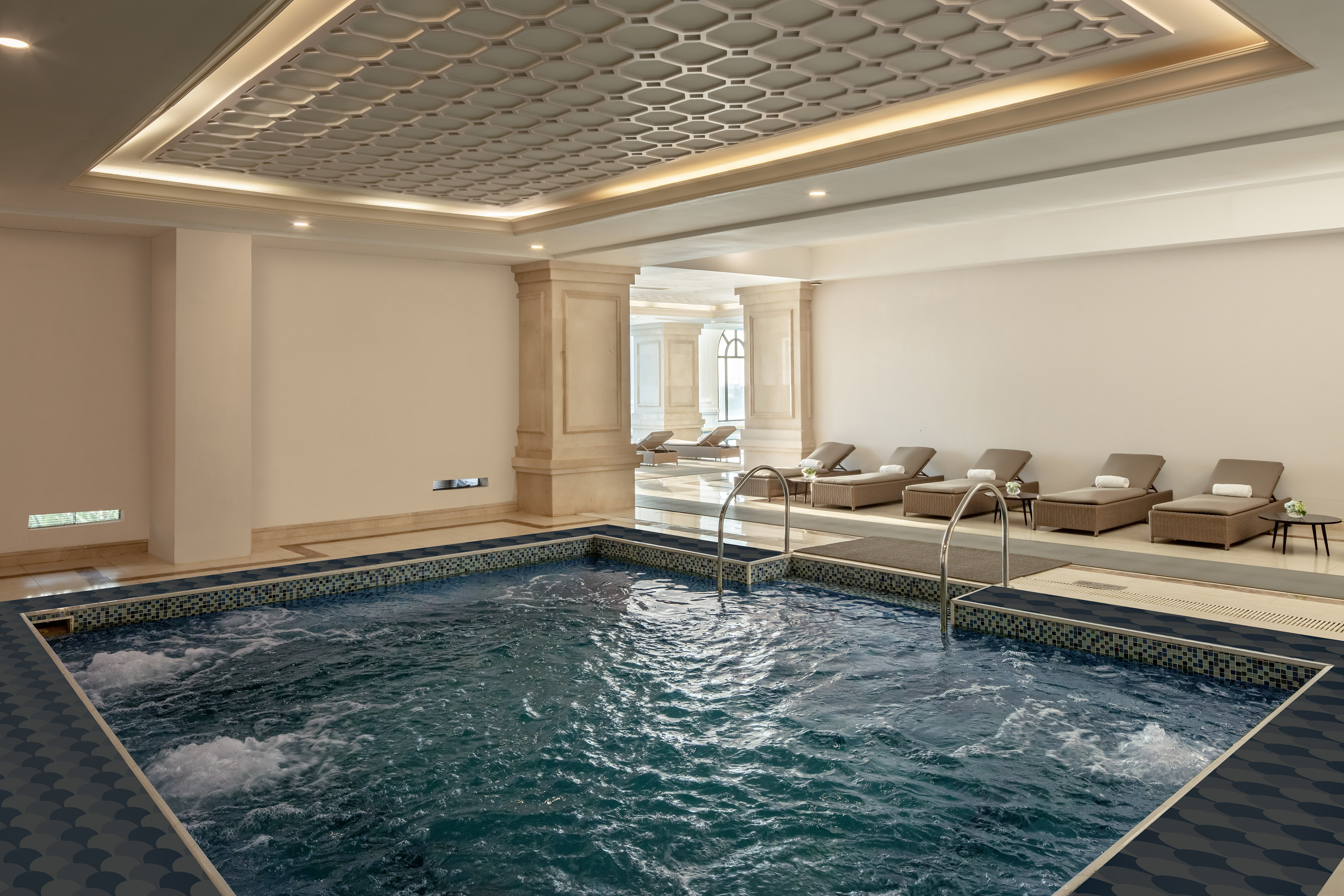 Luxury indoor pool area with bubbling water, lounge chairs, and ornate ceiling.