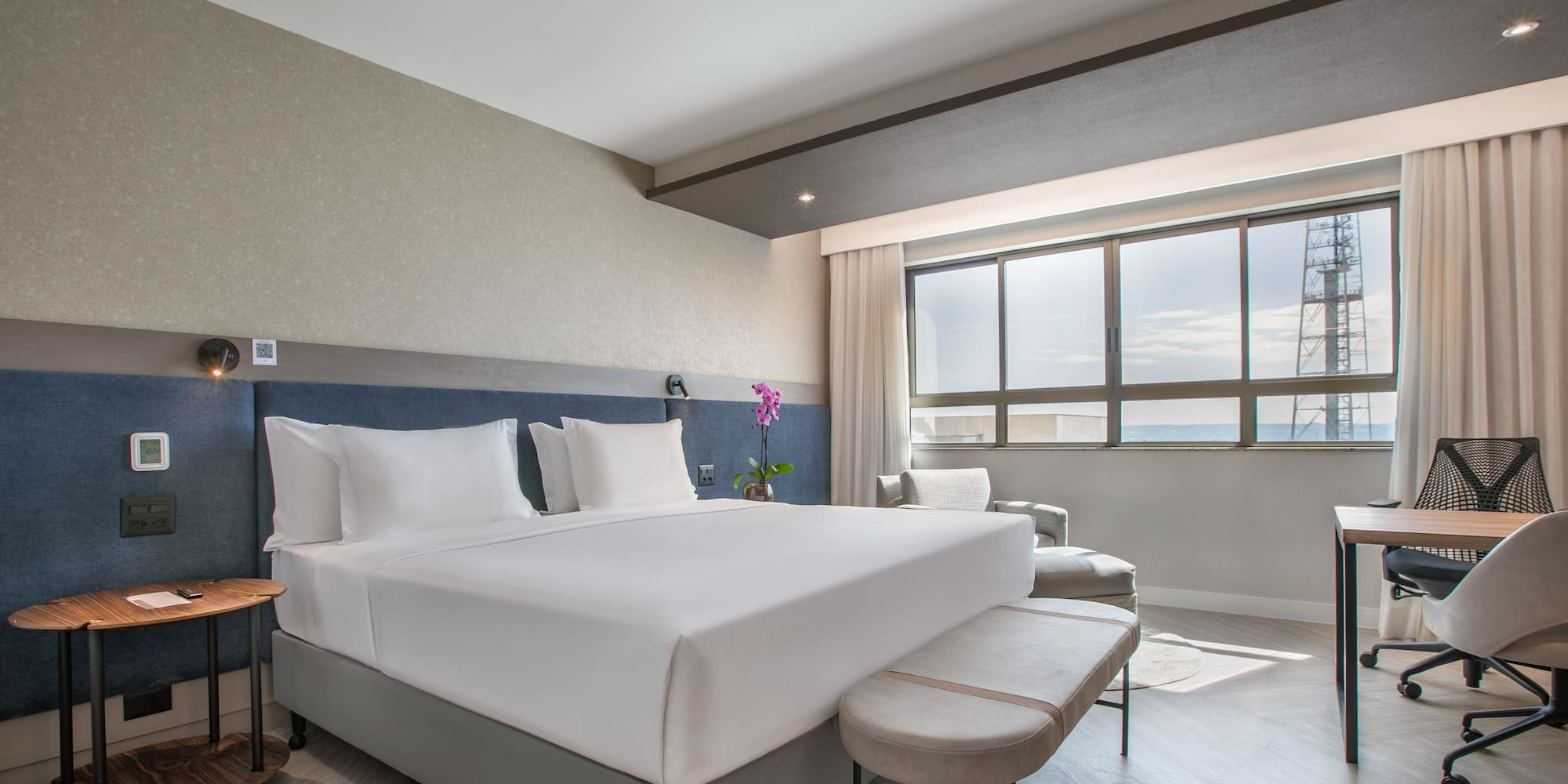 Modern hotel room with king bed, work desk, and large window city view.