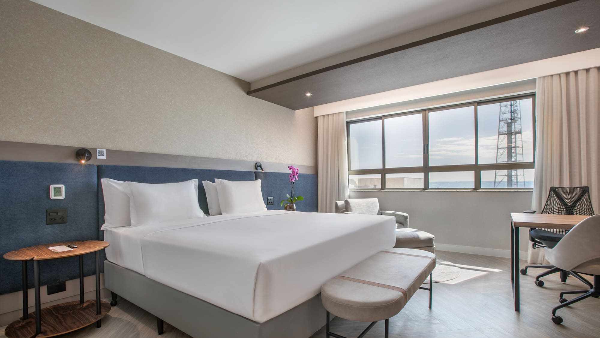 Modern hotel room with king bed, desk, and large window with city view.