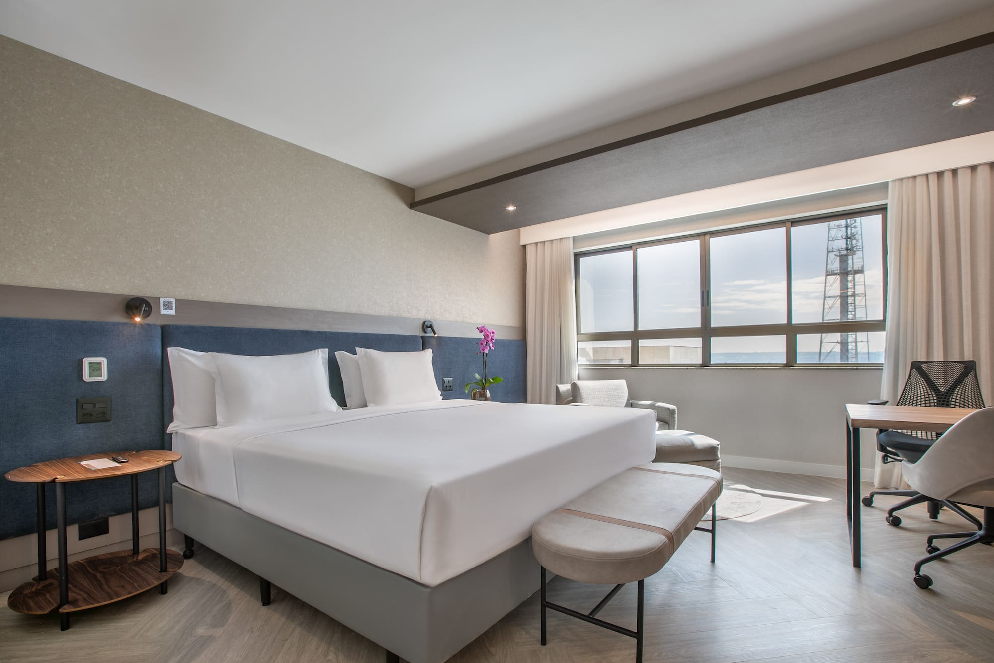 Modern hotel room with king bed, desk, and large window with city view.