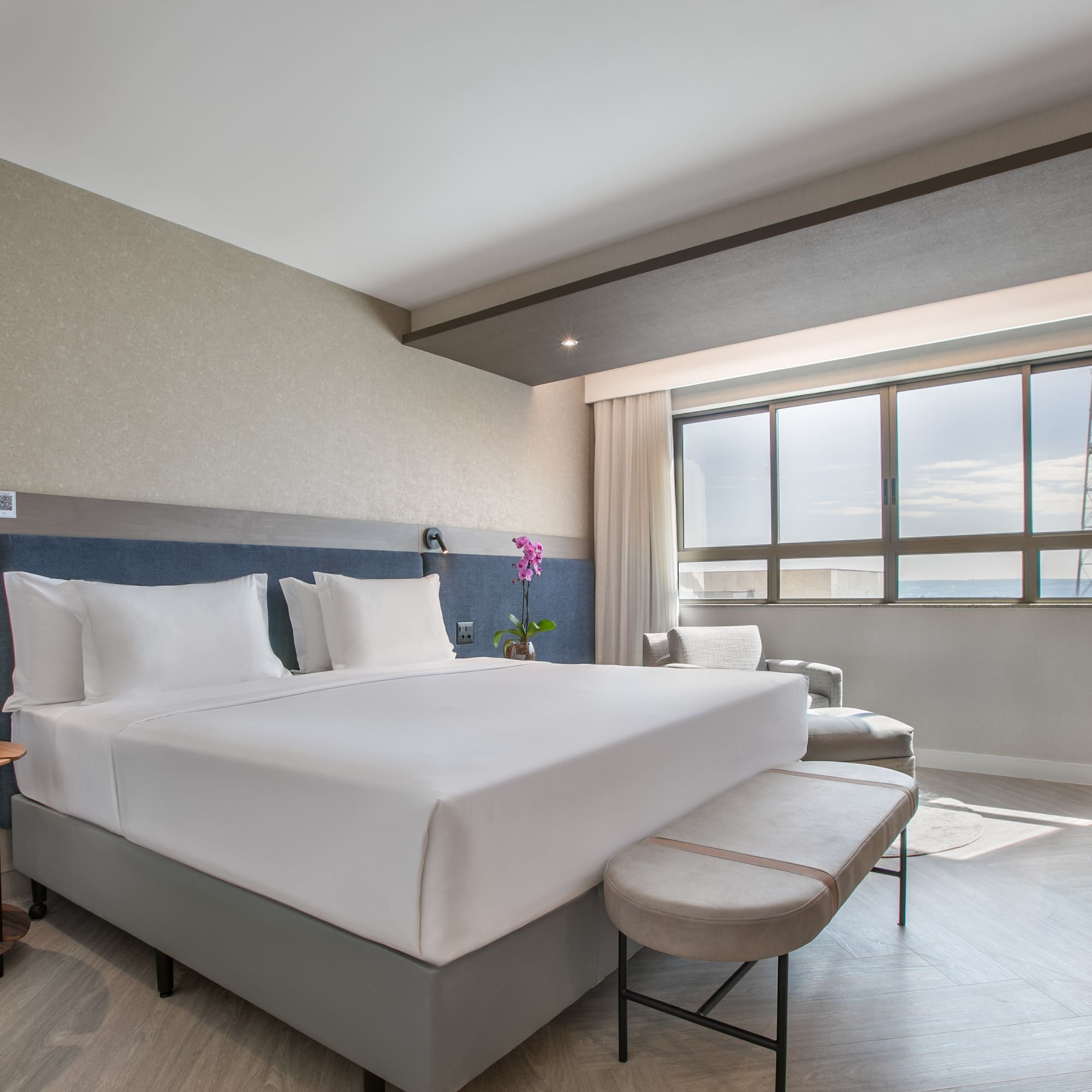 Modern hotel room with king bed, blue headboard, city view window, and workspace.
