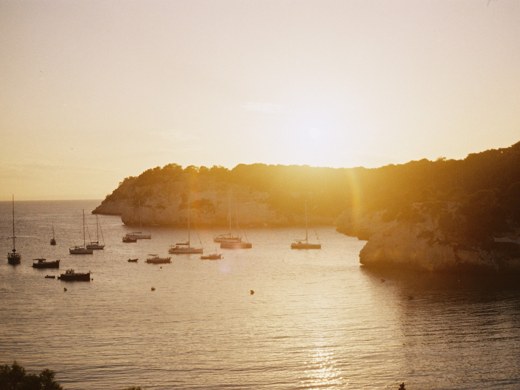 Atardecer dorado en cala con barcos, reflejos en el mar y costa rocosa.